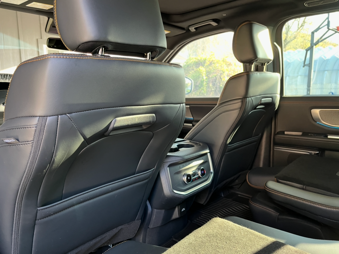 Interior of a vehicle showing the back of the driver's and passenger's seats, with the rear center console, air vents, and window views of trees and a structure with a basketball hoop.