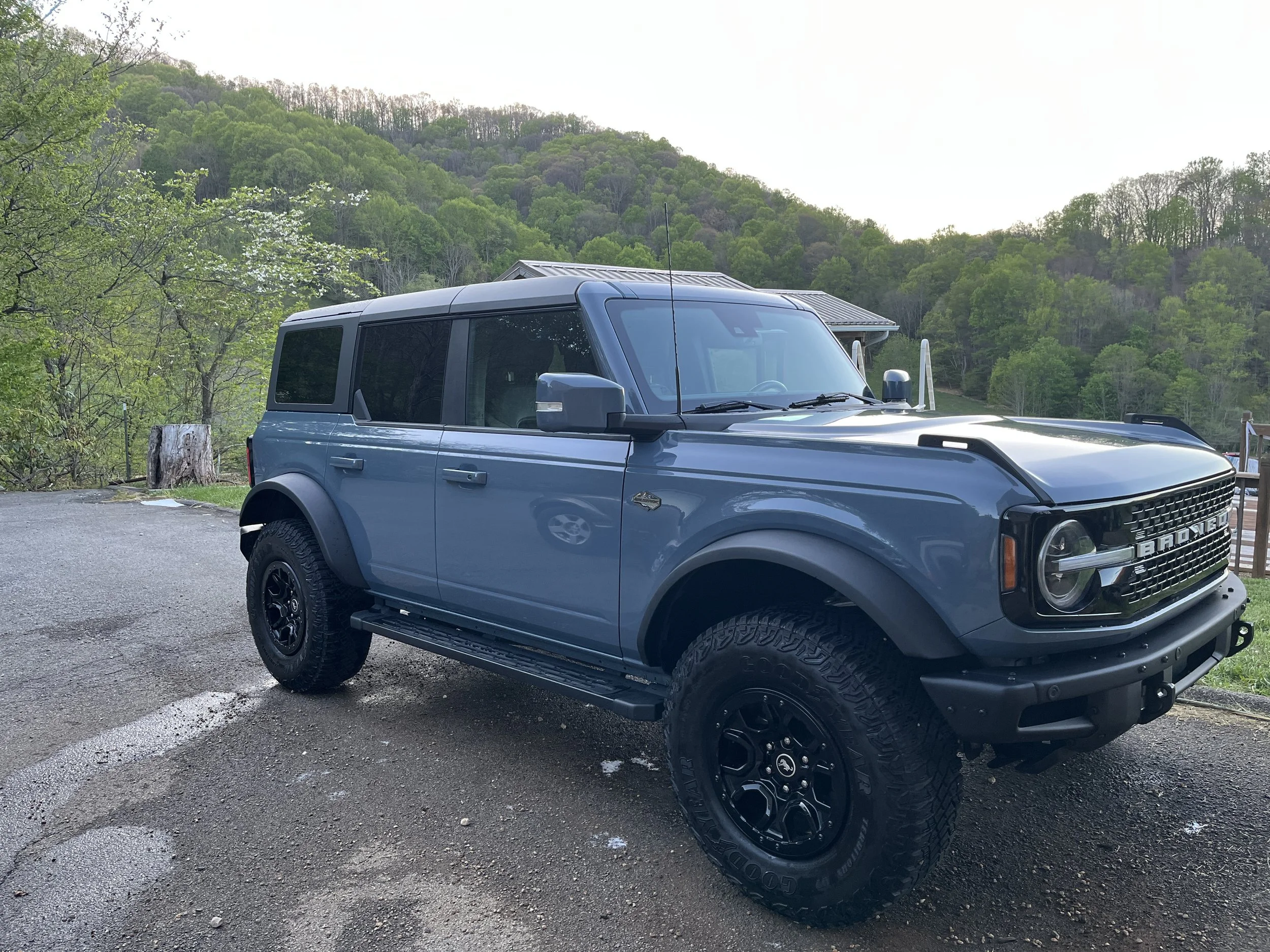 A blue off-road vehicle parked on a gravel surface, with green hills and trees in the background, under a clear sky.