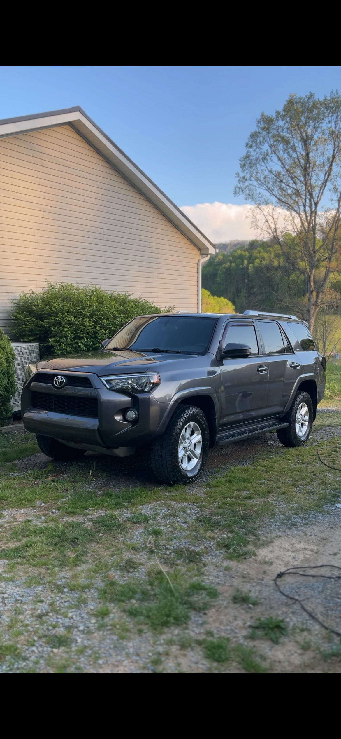 A gray Toyota 4Runner SUV parked on grass and gravel in front of a beige house with siding, green bushes, a tree, and a mountain in the background on a clear day.