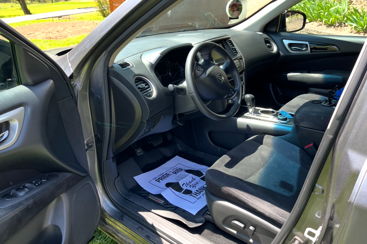 Interior view of a vehicle showing the driver's seat, dashboard, steering wheel, and center console. There are papers on the floor with a message of appreciation. Outside, grass and plants are visible through the open door.