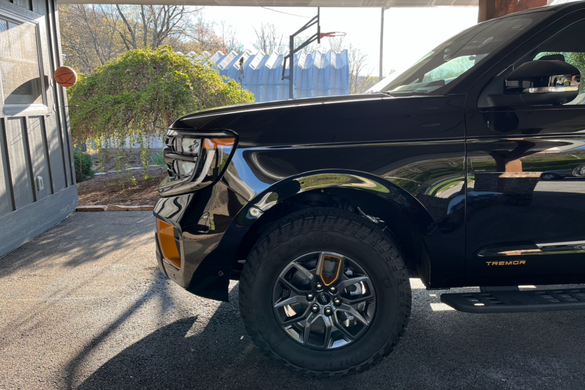Close-up of the front side of a black Ford Tremor pickup truck parked in a driveway with a basketball in a hoop attached to the house wall, a bush, and a blue fence in the background.