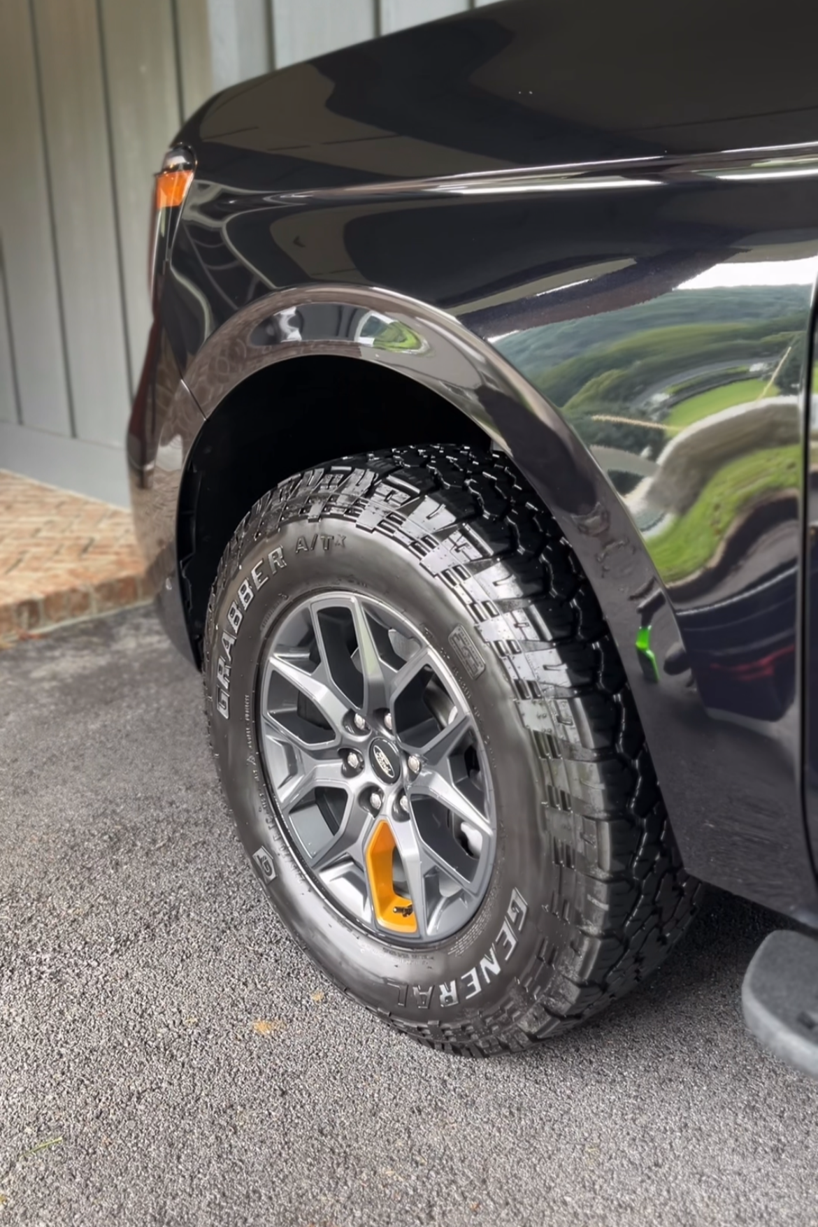 Close-up of the front tire and wheel of a black Ford vehicle, showing rugged off-road tires and a black fender, with a hint of a brick sidewalk and wooden wall in the background.