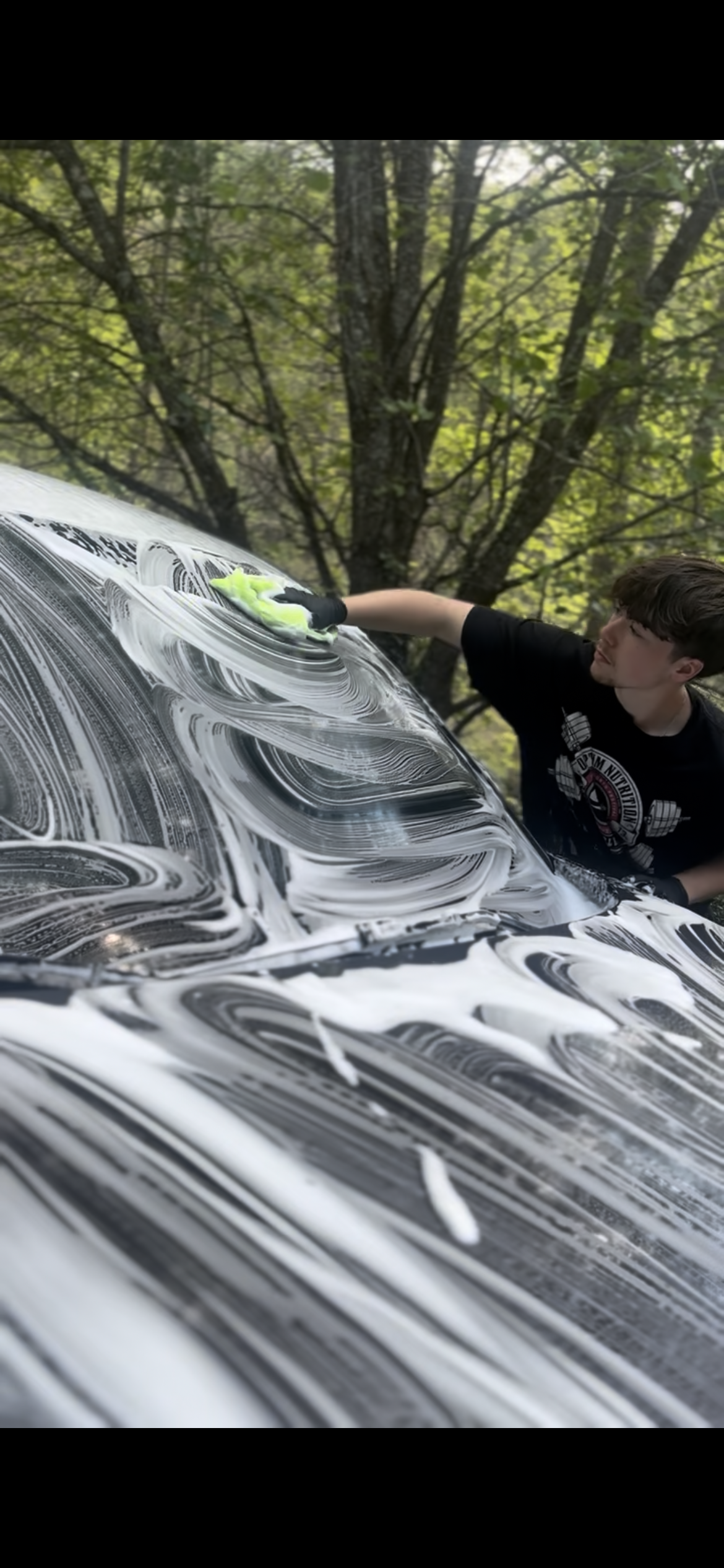 A young man cleaning a car with soap, outdoors in a wooded area.