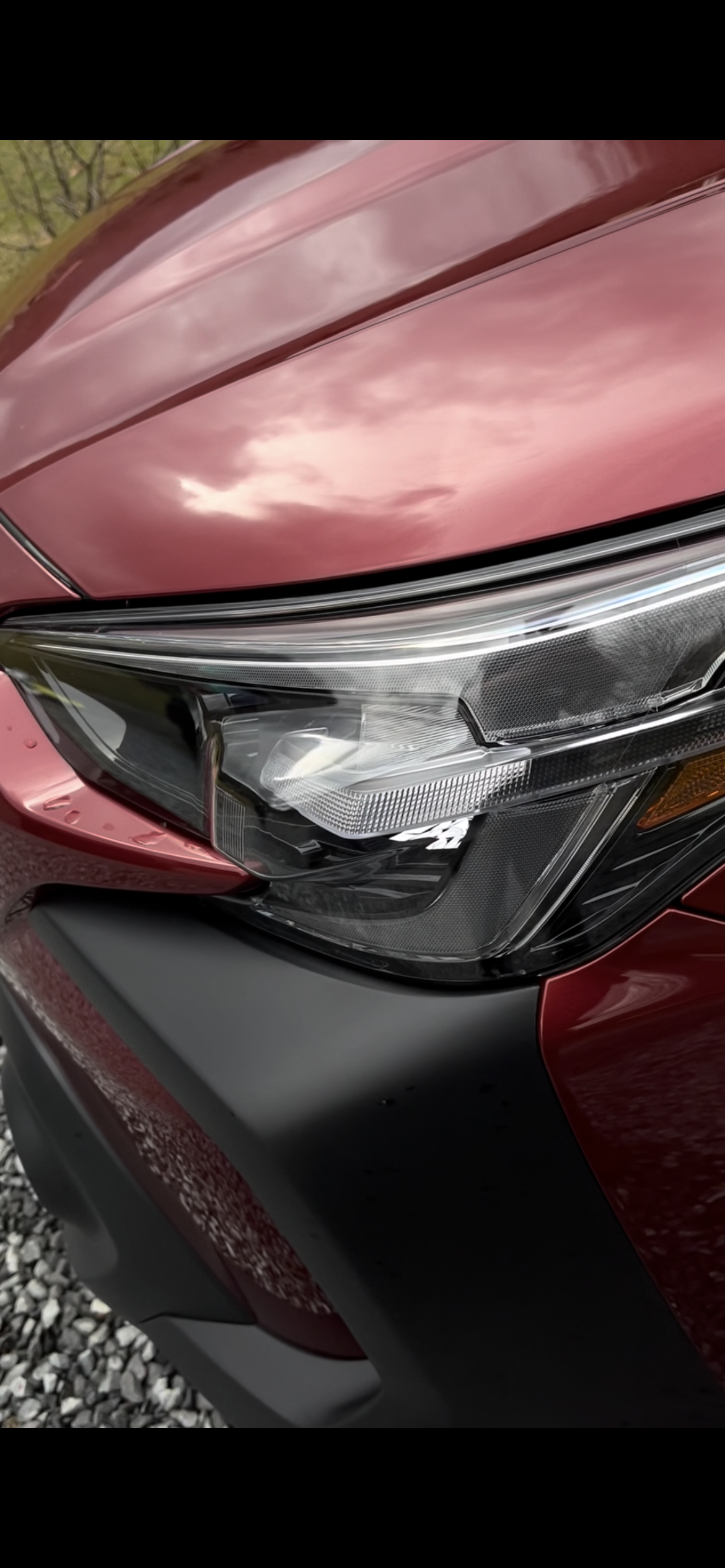 Close-up view of the front right corner of a red car, showing the headlight and part of the hood with reflections of the sky.