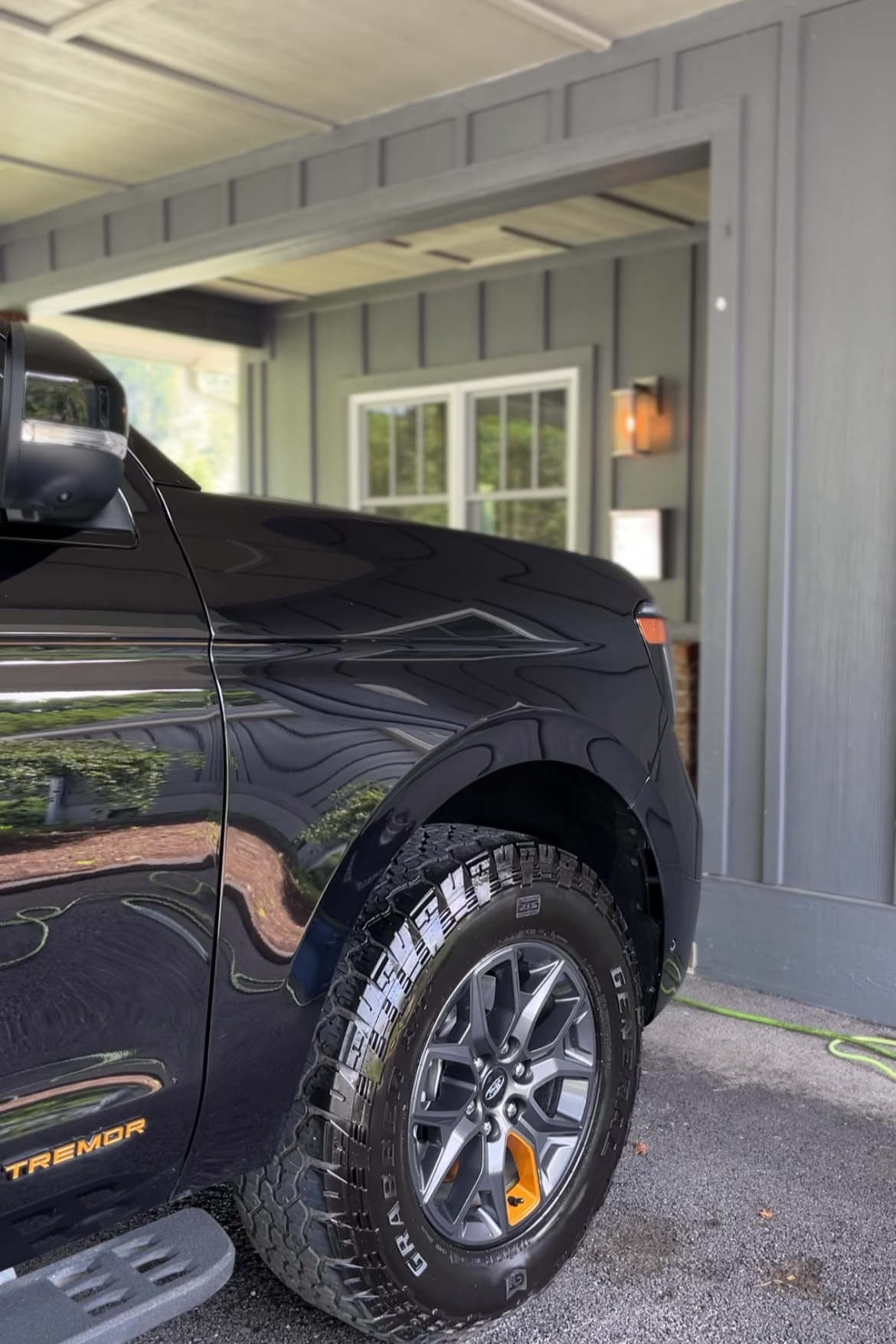 Close-up of the front right corner of a black Ford truck, showing the tire, side step, and part of the door, parked under a covered area with a window and a light fixture on the wall.