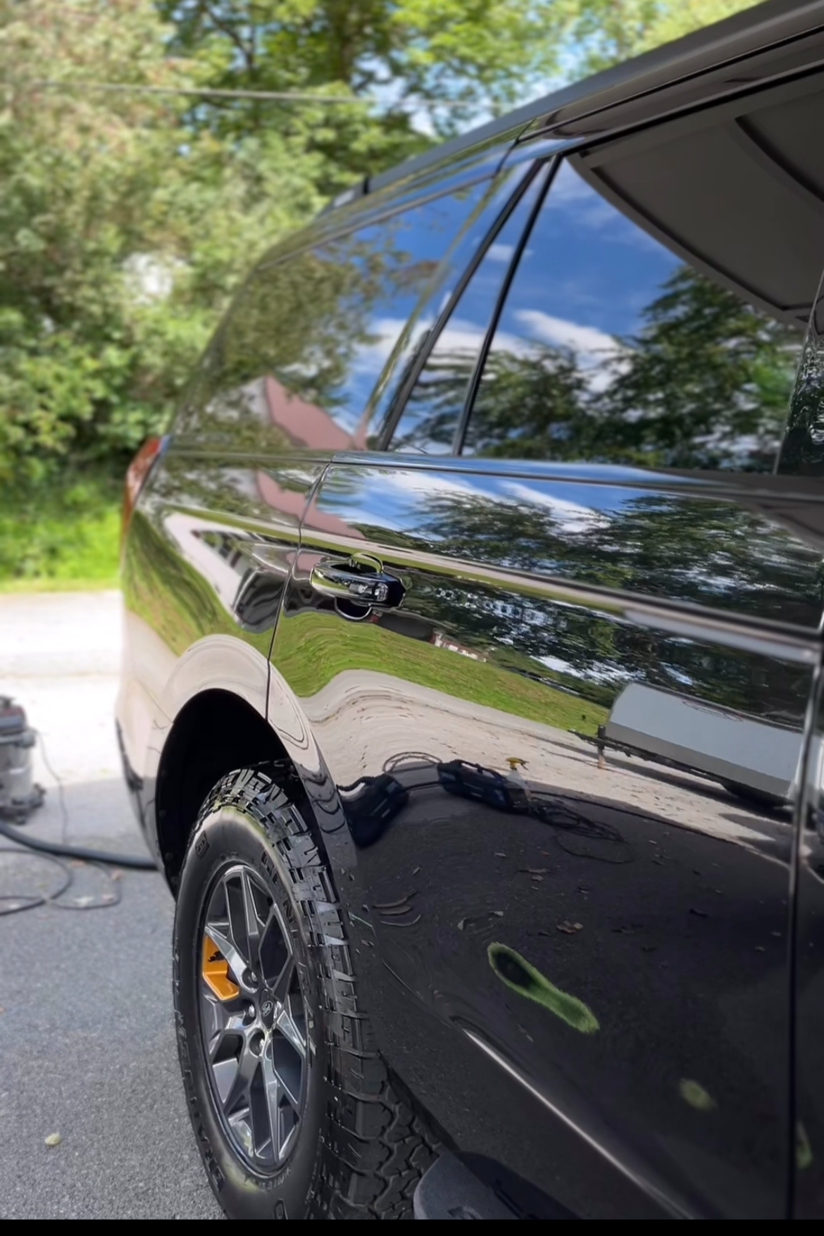 Close-up of a shiny black SUV parked outside, showing reflections of trees and the sky on its surface. The vehicle has off-road tires, and part of the wheel and side panel are visible.