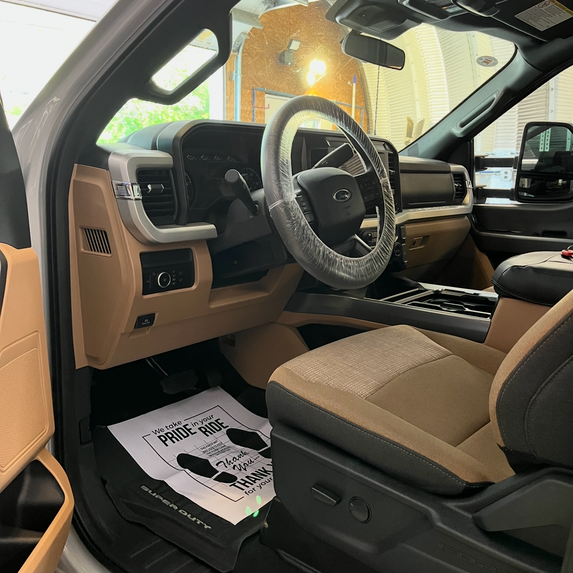 Interior view of a Ford Super Duty truck, showing the dashboard, steering wheel with plastic cover, beige seats, and a paper floor mat that thanks customers for their business.