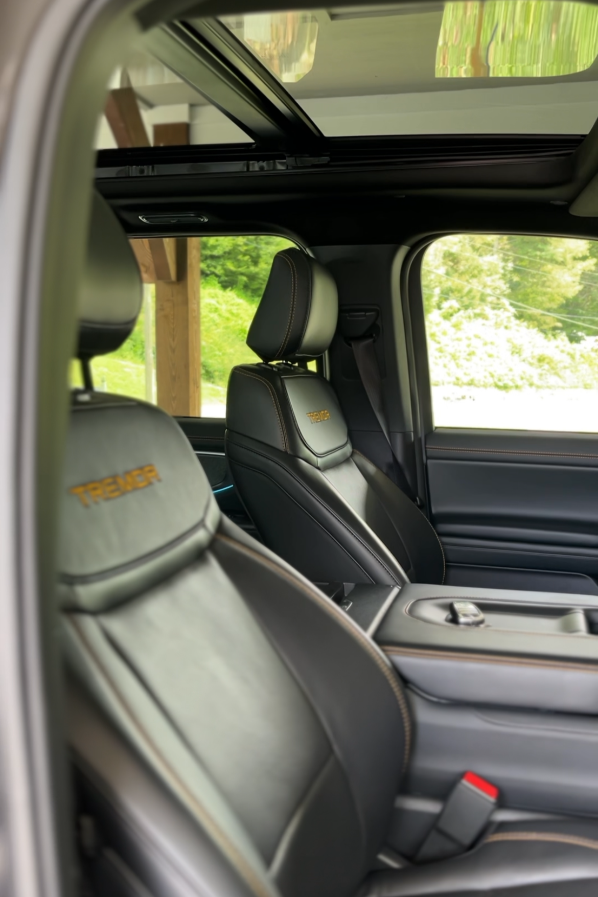 Interior of a vehicle showcasing black leather seats with yellow stitching and the word 'TREK' embroidered on the headrests, a view through the window of green trees and a wooden structure.