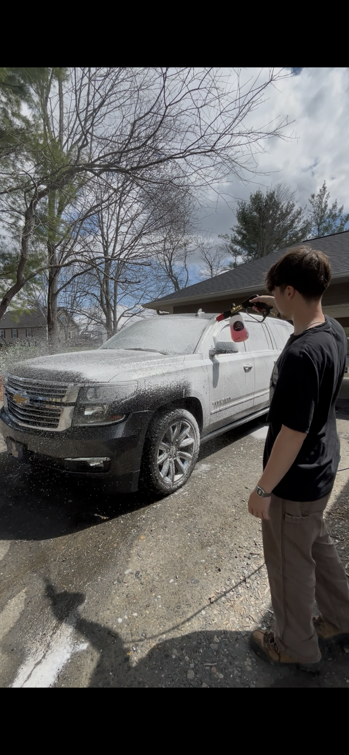 A young man in black shirt and khaki pants washing a gray SUV with a foam cannon. The vehicle is parked outdoors near a house, with trees in the background and a partly cloudy sky.