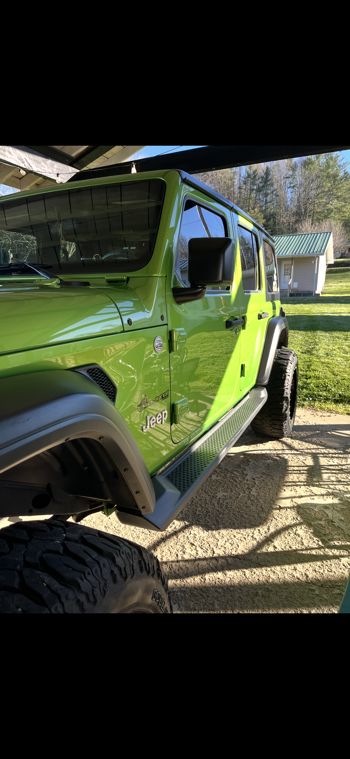 A bright green Jeep Wrangler parked outside on a sunny day, with trees and a small building in the background.