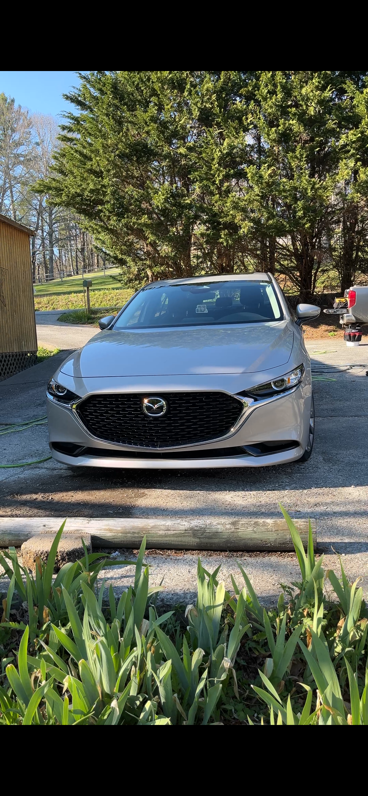 Silver Mazda sedan parked outdoors with greenery, trees, and a wooden structure in the background.