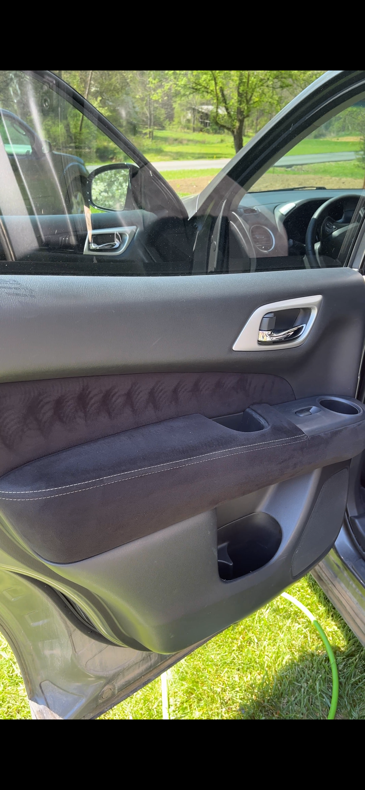 Close-up of the inside of a car door, showing the window, door handle, armrest, and storage compartment, with a green lawn and trees visible outside.