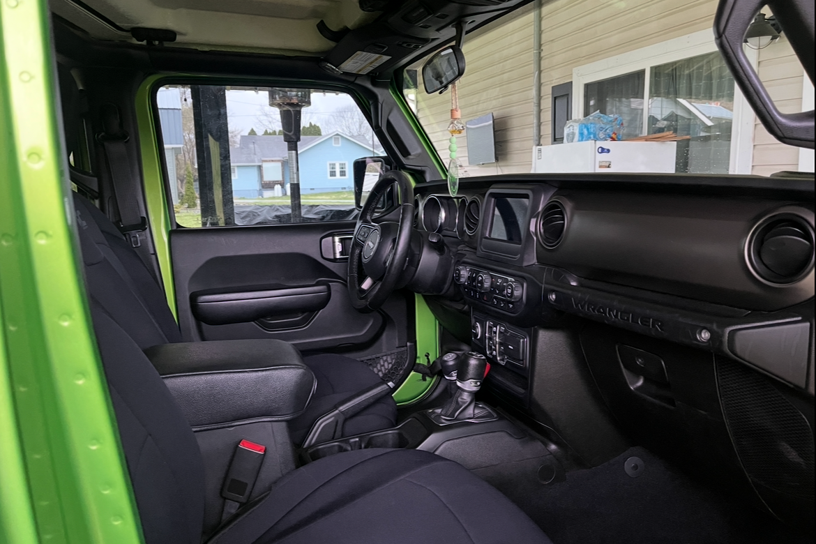 The interior of a green Jeep Wrangler showing the driver’s seat, dashboard, steering wheel, and front passenger seat, with a glimpse of the outside through the windshield.