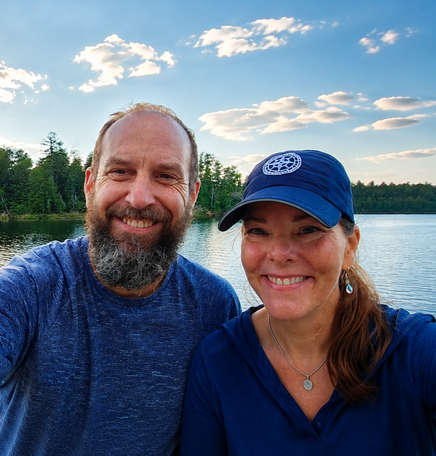 A happy man and woman smiling at the camera near a lake with trees and a blue sky with clouds in the background.
