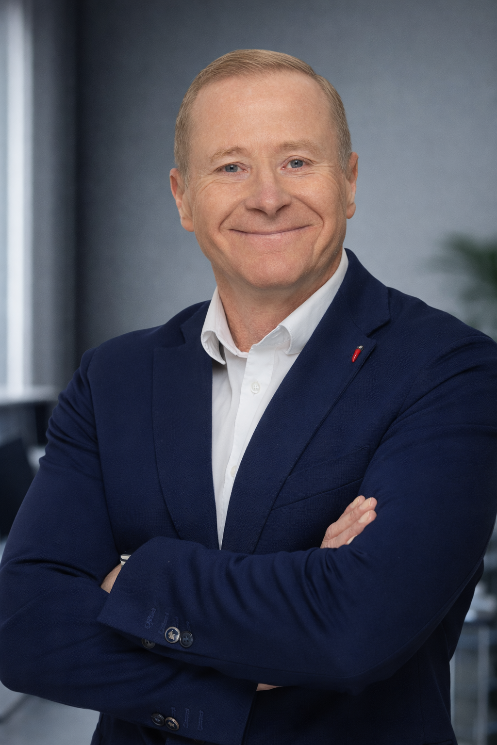 A middle-aged man with short blonde hair, wearing a navy blue suit and white shirt, smiling with arms crossed in an office setting.