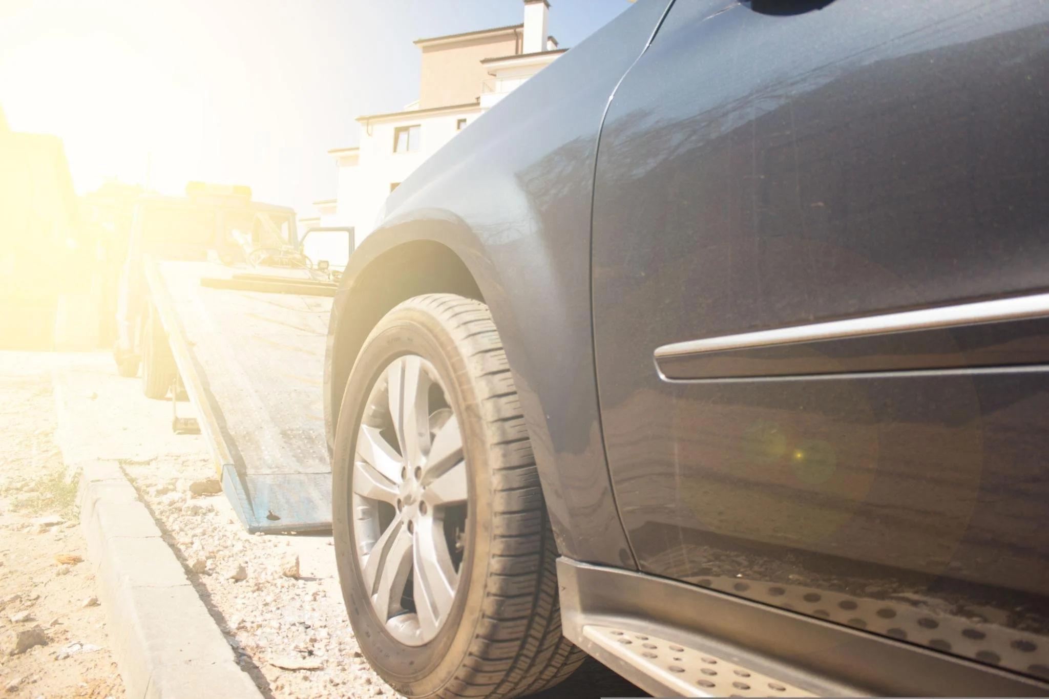 Close-up of the side of a gray luxury car, highlighting its wheel and part of the door, parked on a sunlit street with a building in the background.