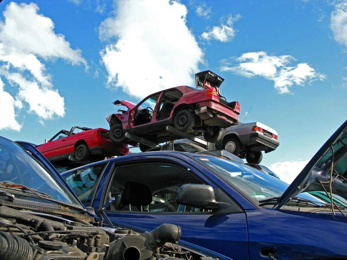 Stacked cars in a salvage yard with a blue car in the foreground, a red car on top of other vehicles, and a cloudy sky.