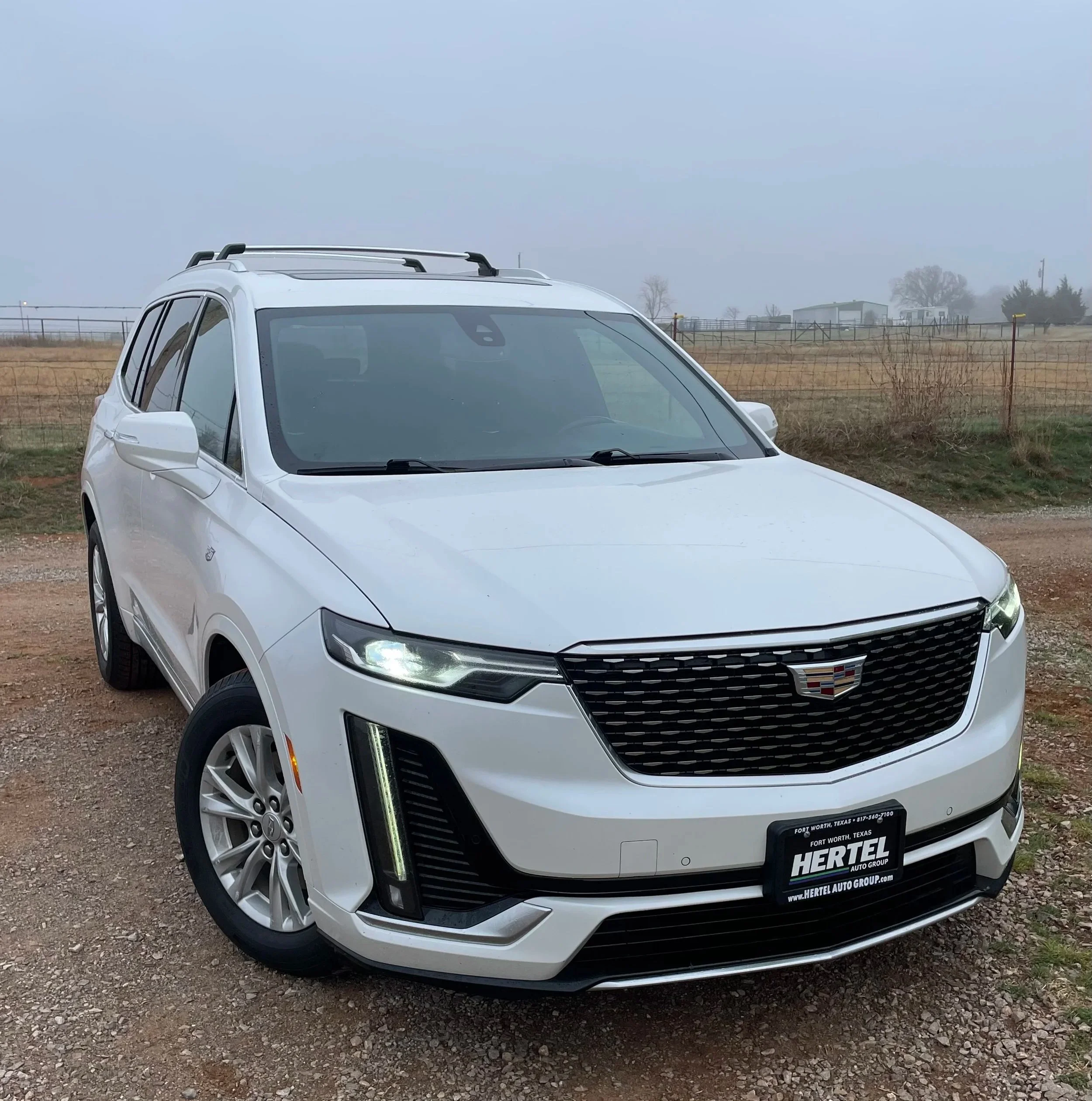 Front view of a white Cadillac SUV parked on a dirt patch with a rural background, including a fence, open fields, and overcast sky.