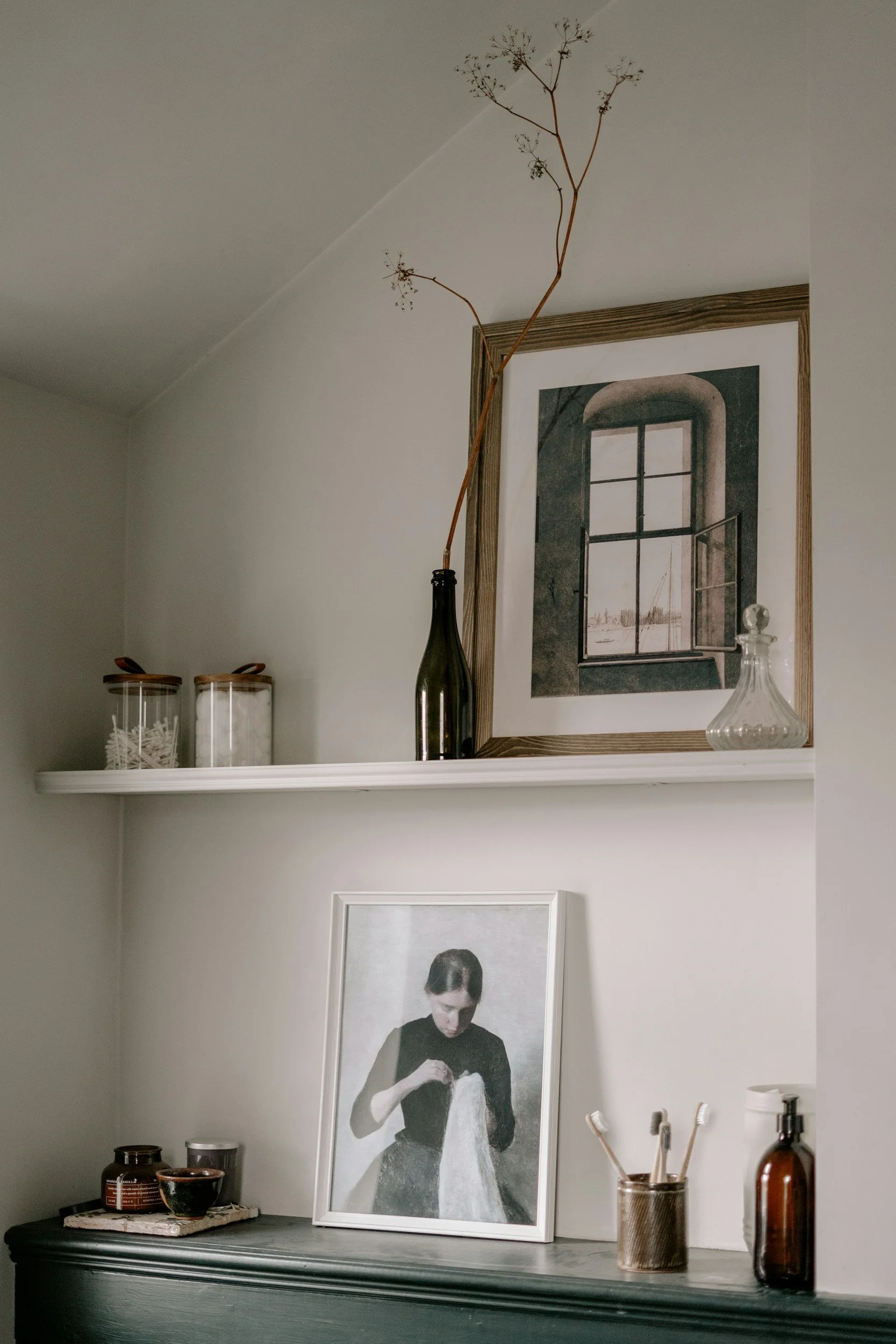 Decorative shelf with jars, a framed photo of a woman holding fabric, a framed window photograph, and a vase with a dried branch.