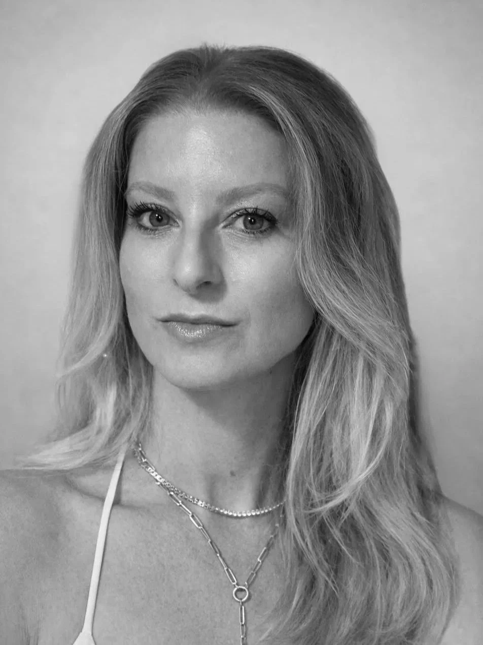 Black and white portrait of a woman with wavy, shoulder-length hair, wearing a layered necklace, and looking directly at the camera.