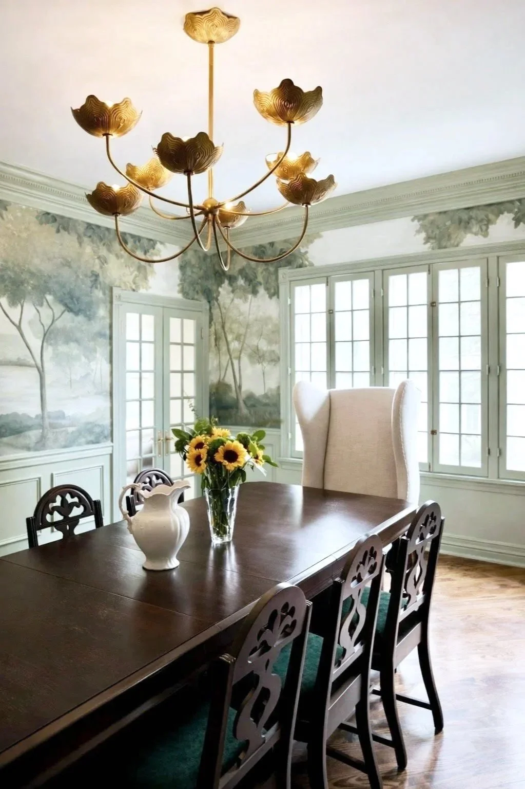 A dining room with a dark wooden table, a vase of sunflowers, and a white pitcher on it. Six black chairs with decorative backs surround the table. The room has large windows and a wall mural of a landscape. A chandelier with gold flower-shaped shades hangs from the ceiling.