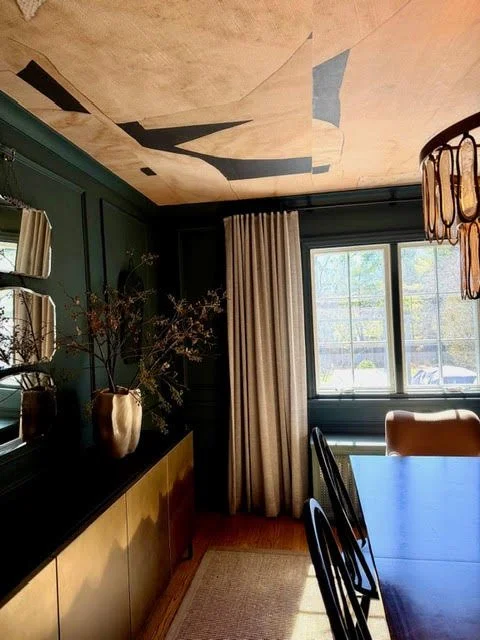 Interior view of a dining room with a dark green wall, beige curtains, a large window, and a wooden dining table with black chairs. A ceiling with abstract painted design, a decorative hanging light fixture, and a sideboard with vases and branches are also visible.