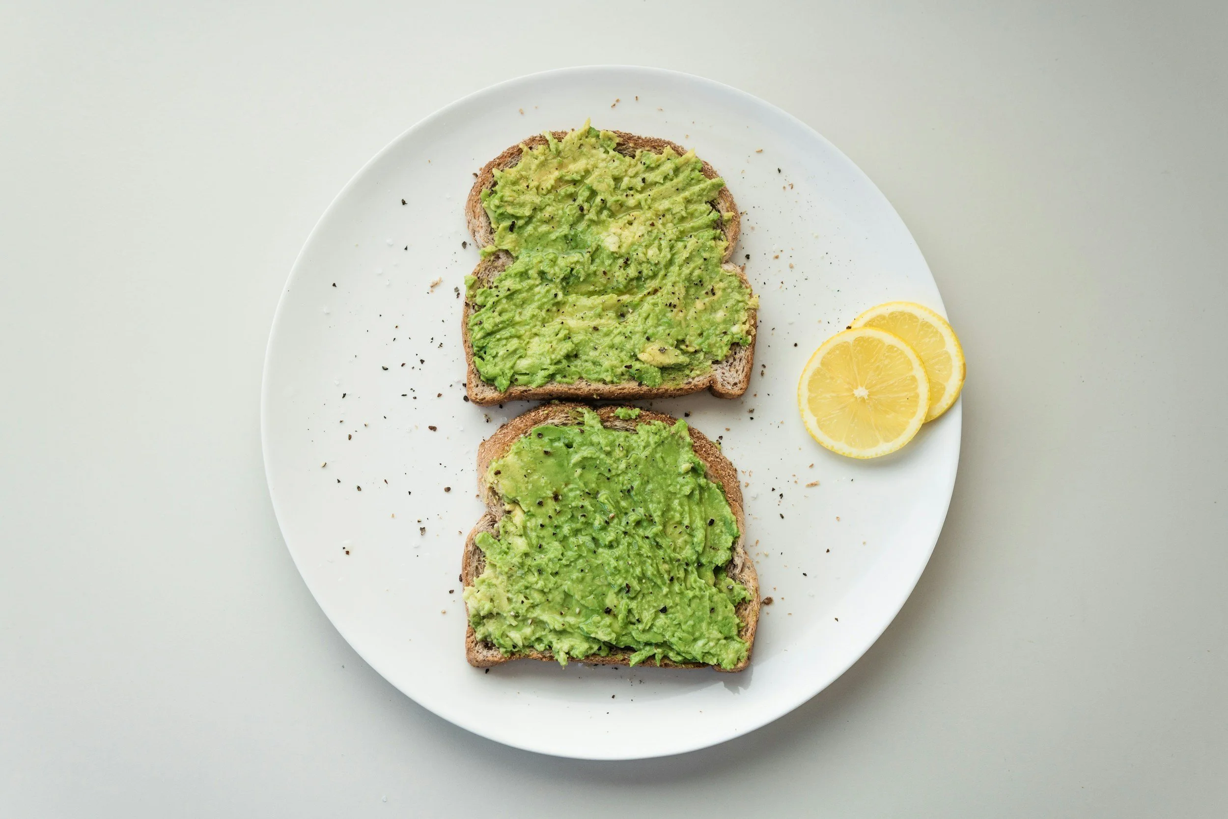 Three slices of toast with mashed avocado, sprinkled with black pepper on a white plate, with two lemon slices on the side.