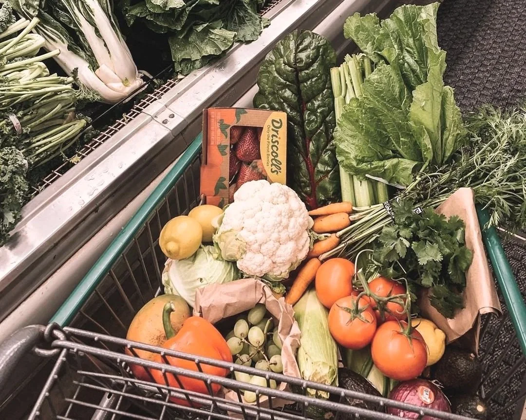 Cart filled with fresh vegetables including cauliflower, tomatoes, carrots, green onions, lettuce, cilantro, strawberries, and other produce, in a grocery store.