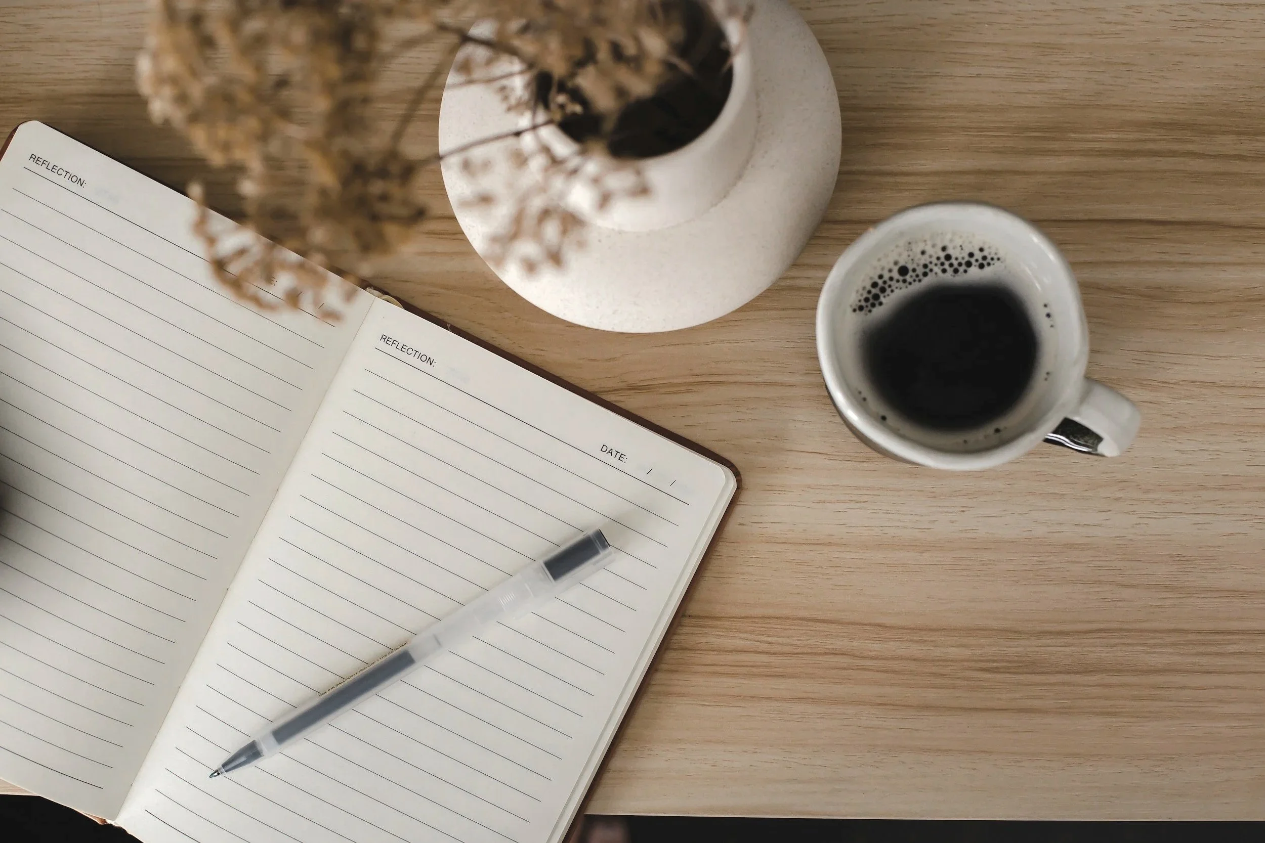 An open notebook with lined pages on a wooden desk, a silver pen placed on it, a black cup of coffee, and a small white vase with dried plants.
