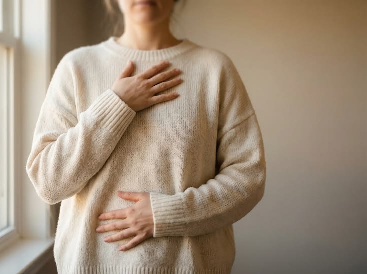 Person wearing a cream-colored sweater, holding their hand on their chest and stomach, standing indoors near a window.