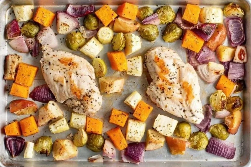 Baked chicken breast fillets on a tray with roasted vegetables, including Brussels sprouts, sweet potatoes, red onions, and parsnips.