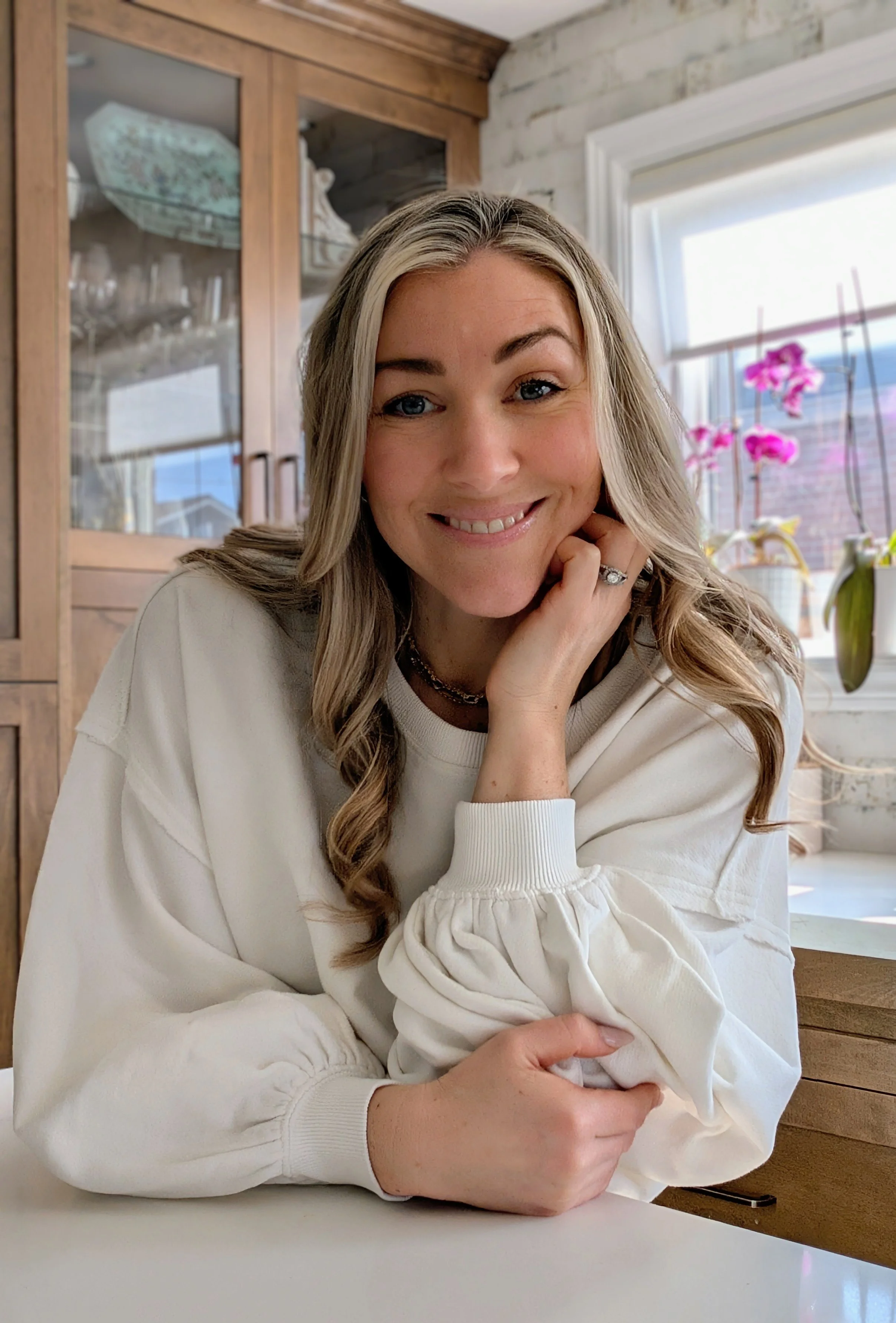 A woman with long wavy blonde hair, wearing a white sweatshirt, is smiling and resting her chin on her hand. She is sitting at a white table in a bright room with a window and pink flowers in the background.