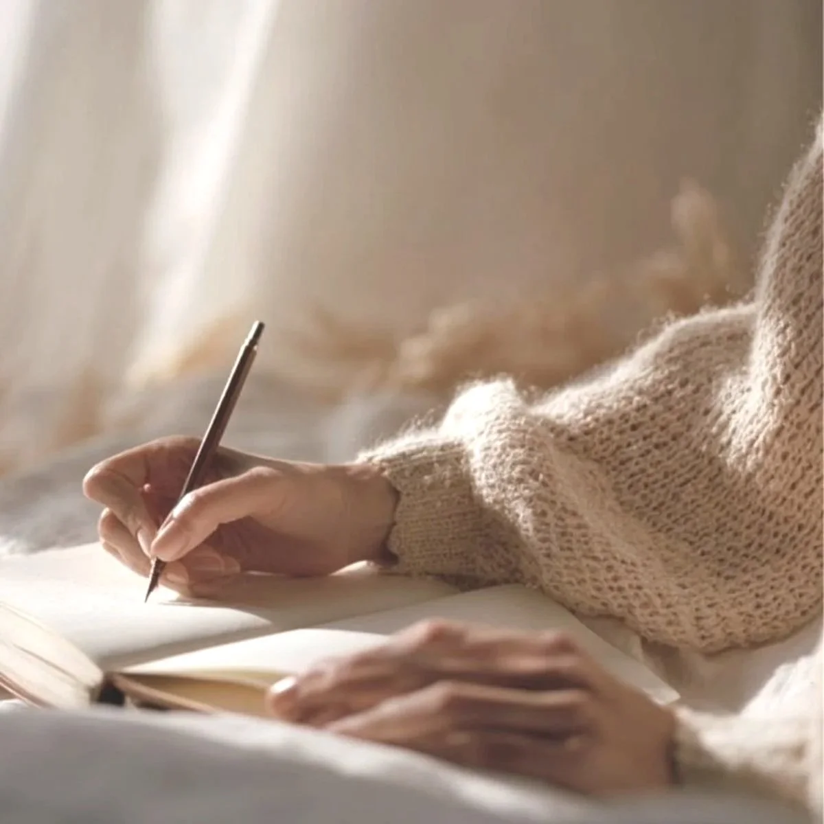 Person in a cozy beige sweater writing in a notebook with a pen indoors.