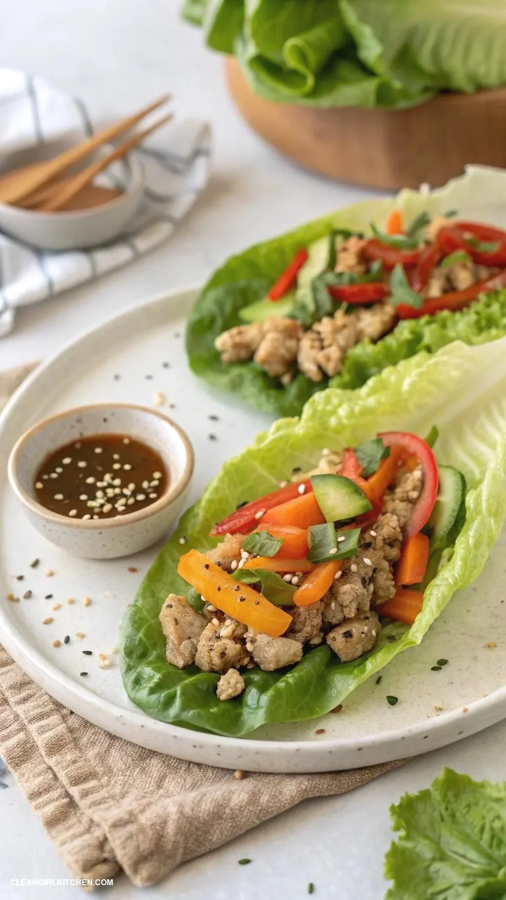 Fresh lettuce wraps filled with cooked ground chicken, red and orange bell peppers, cucumber slices, and green onions, served with a side of soy sauce topped with sesame seeds.