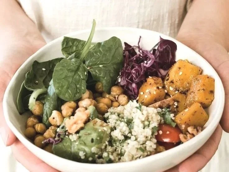 A white bowl filled with a variety of fresh salad greens, chickpeas, roasted sweet potatoes, crumbled cheese, and cherry tomato slices.