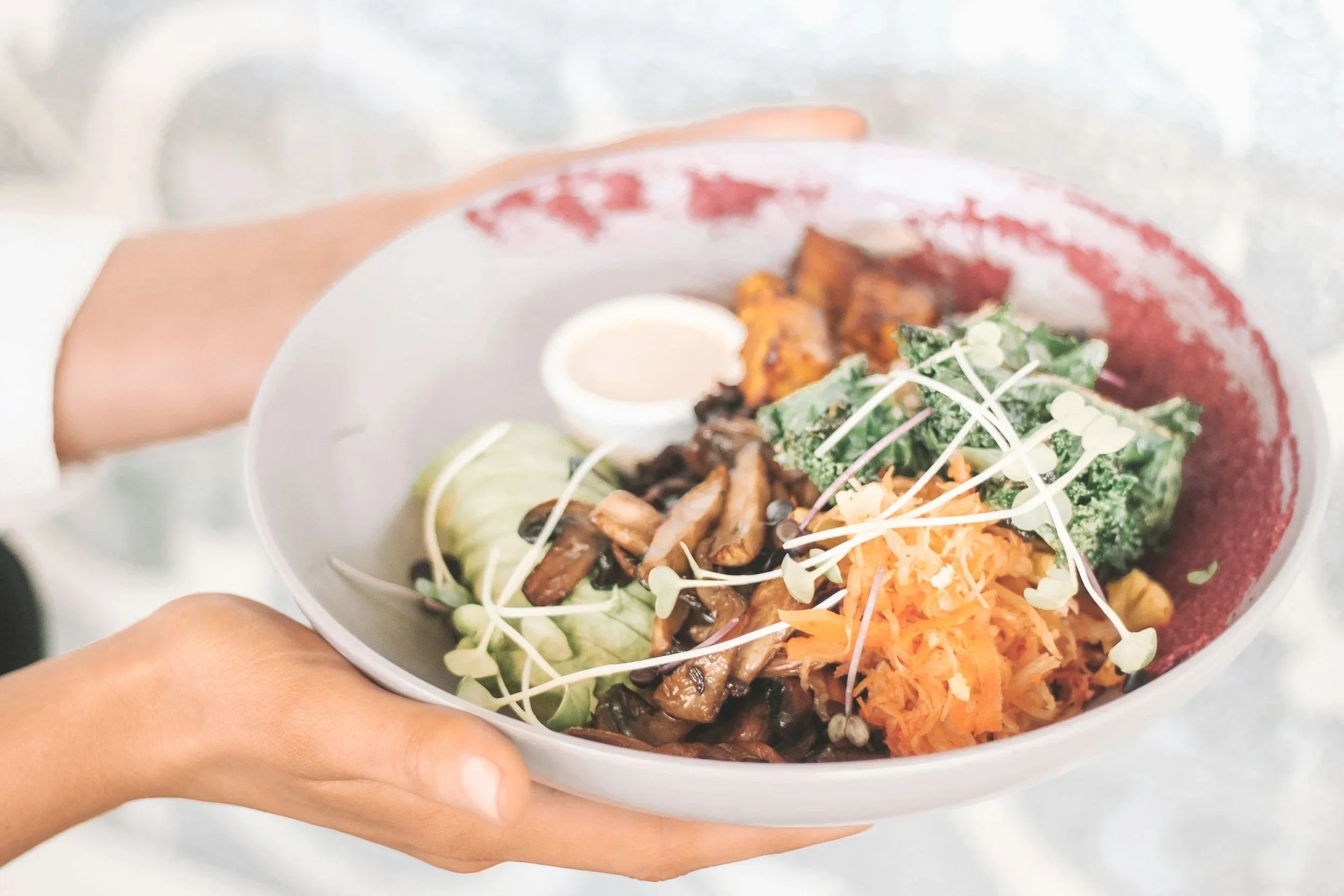 A hand holding a bowl filled with shredded carrots, kale, microgreens, grilled vegetables, and a boiled egg, with a red sauce or dip on the rim of the bowl.