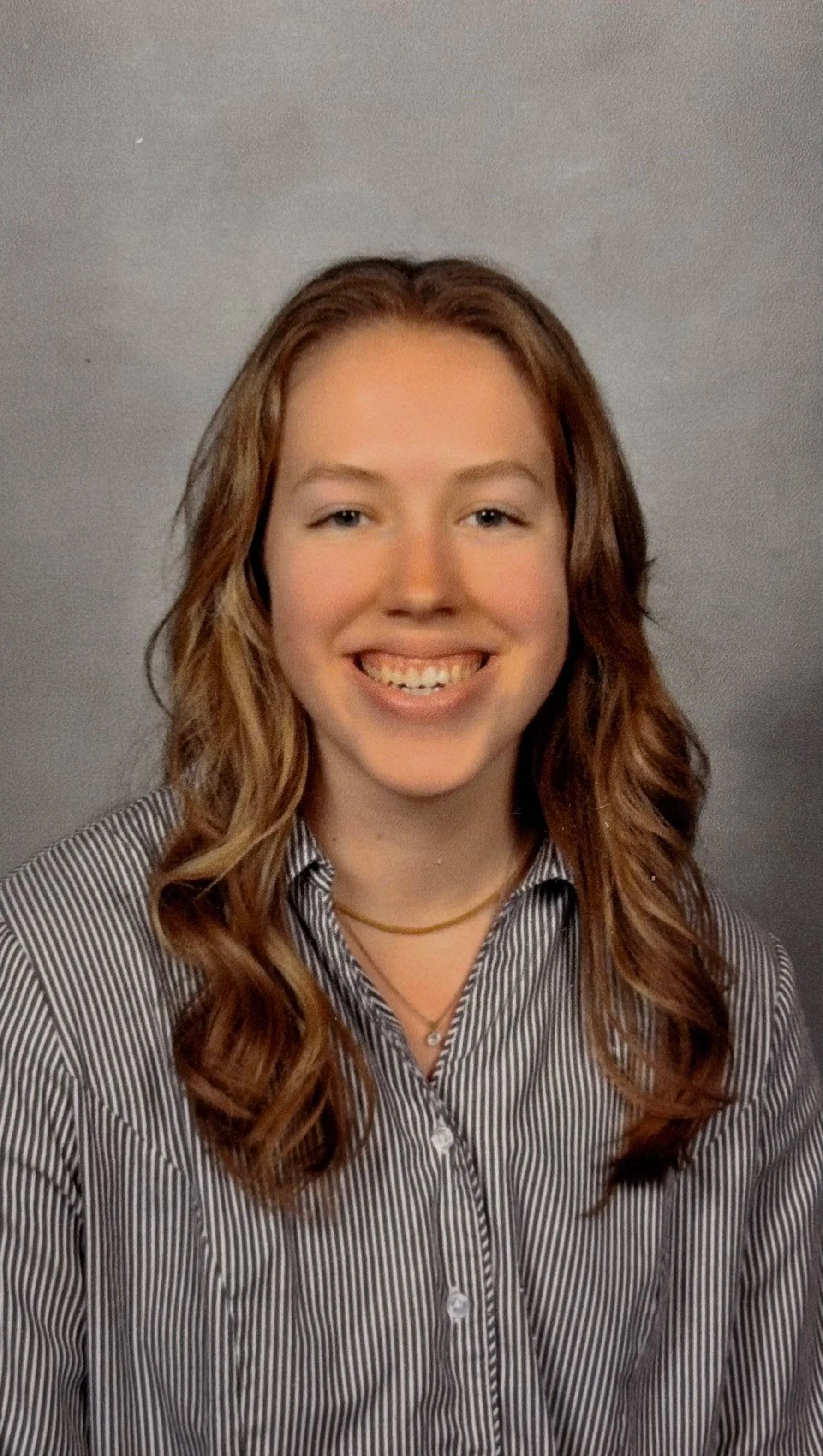 A young woman with long, wavy auburn hair smiling at the camera, wearing a black and white striped button-up shirt and layered necklaces, against a plain gray background.