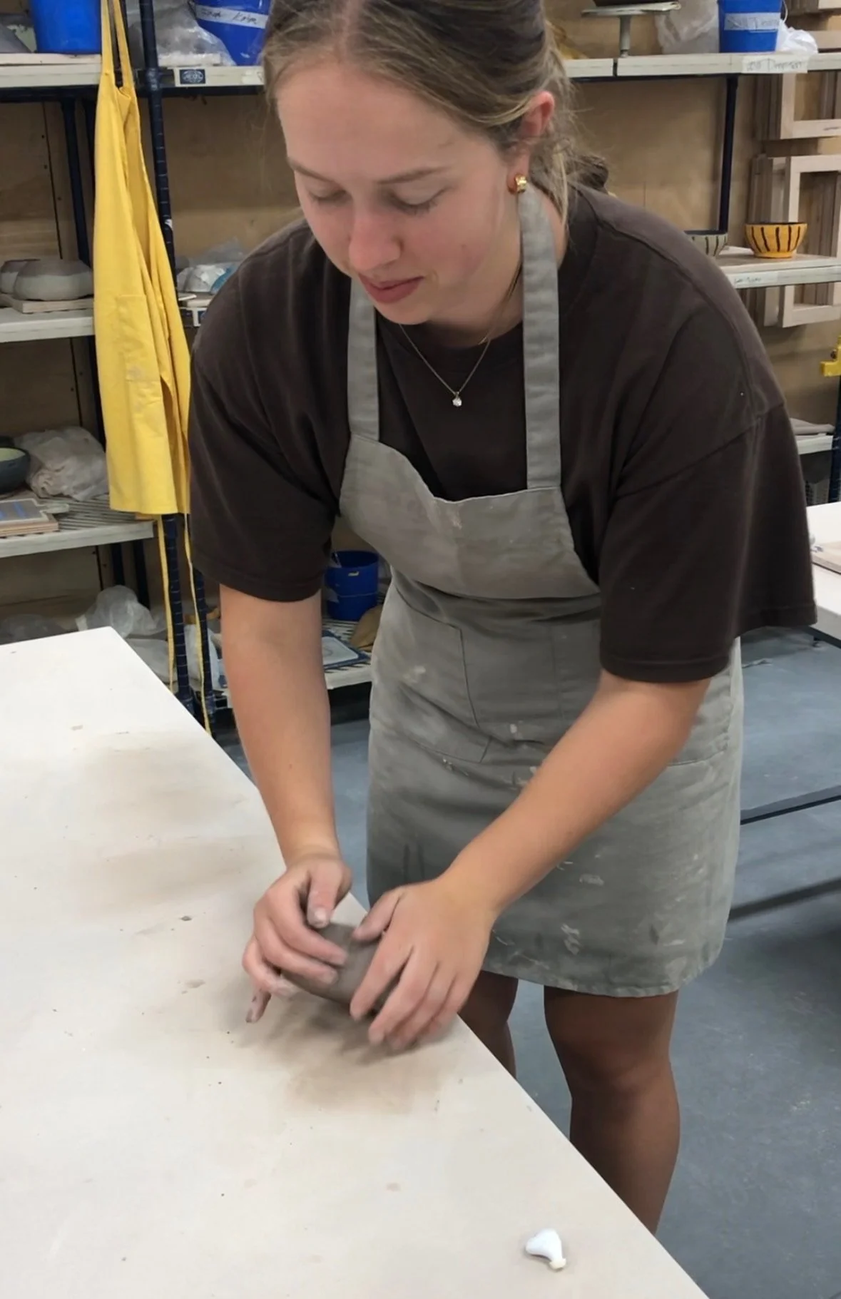 A woman in a gray apron rolling clay on a table in a pottery studio.