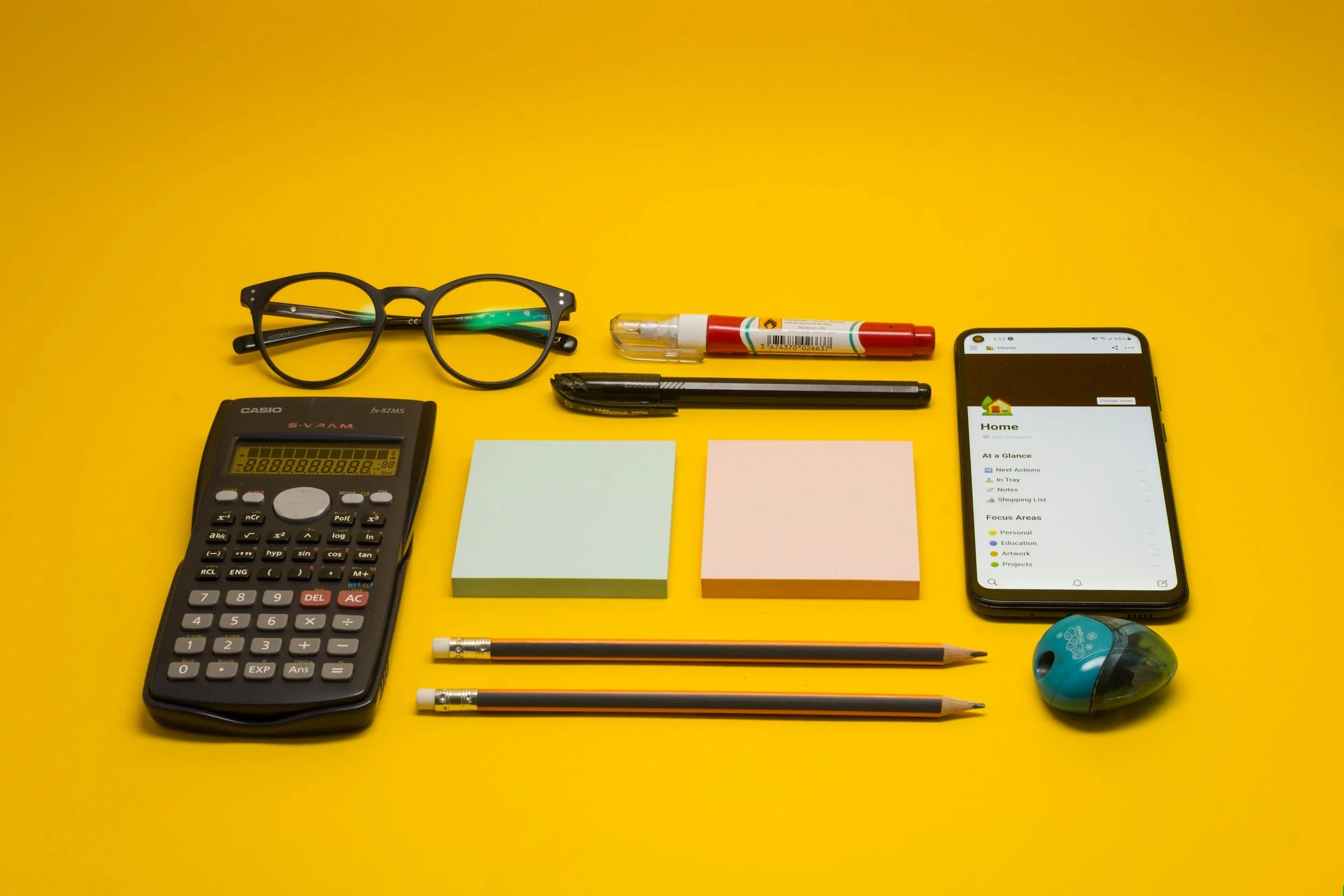 Desk with yellow background, items include black glasses, a red and clear spray bottle, black pen, sticky notes in blue and pink, a calculator, a pencil, a smartphone displaying a home organization app, and blue wireless earbuds.