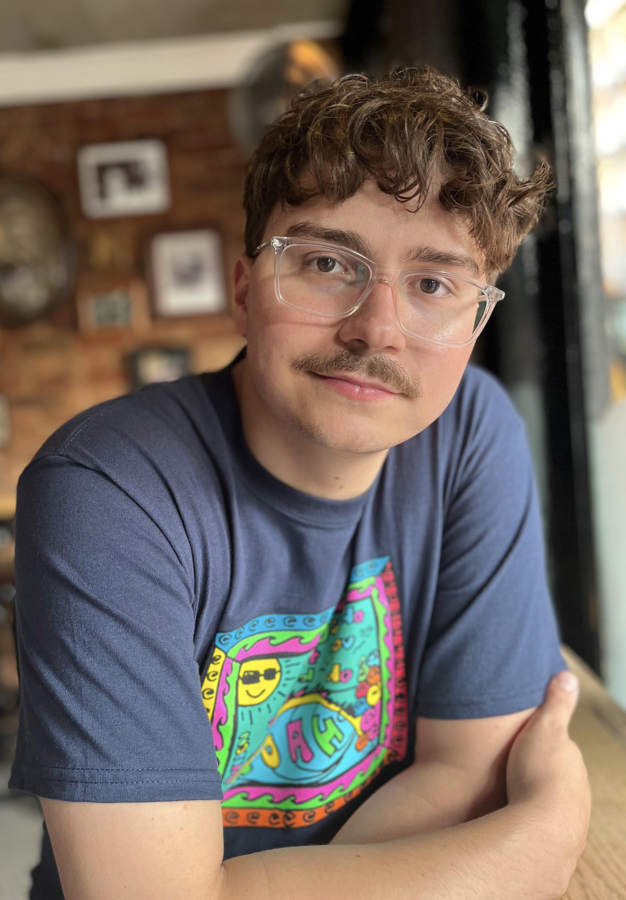 A young man with curly brown hair, glasses, and a mustache is smiling at the camera. He is wearing a dark T-shirt with a colorful graphic on it. He is sitting at a table with his arms crossed. The background shows framed pictures on a brick wall and a window with black curtains, indicating he is inside a cozy café or restaurant.