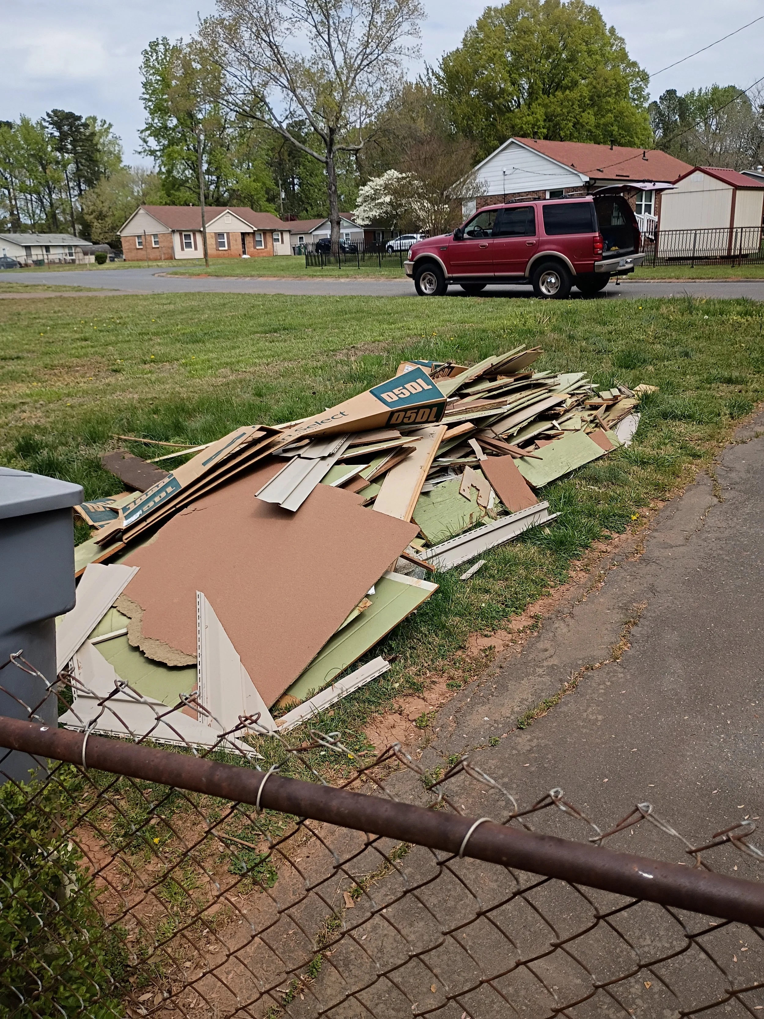 A pile of discarded drywall and cardboard boxes in a grassy area next to a chain-link fence, with a residential street and houses in the background under a cloudy sky.