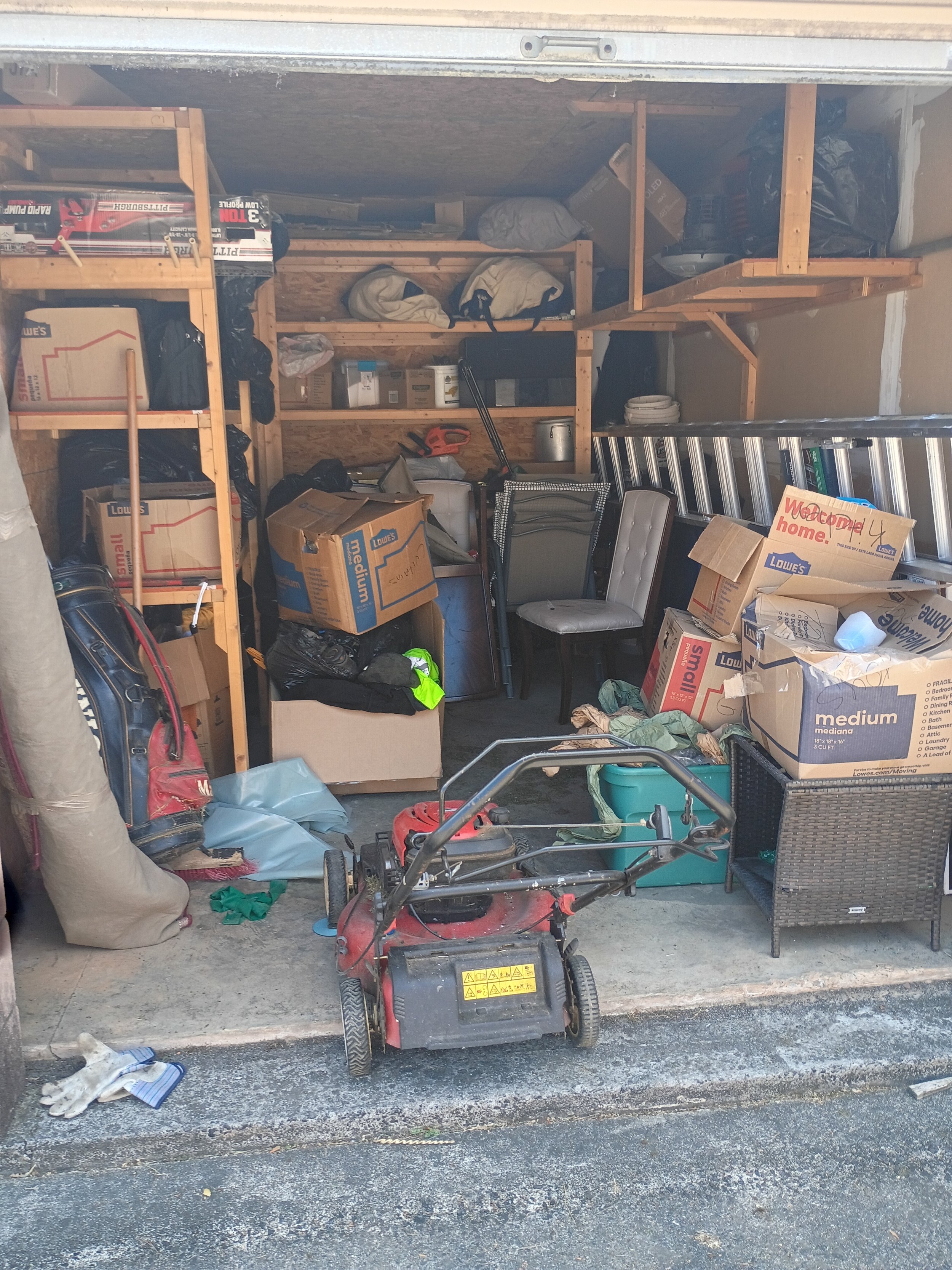 A cluttered garage with shelves, boxes, a lawnmower, and various stored items. Before Hallelujah Hauling cleaned it.