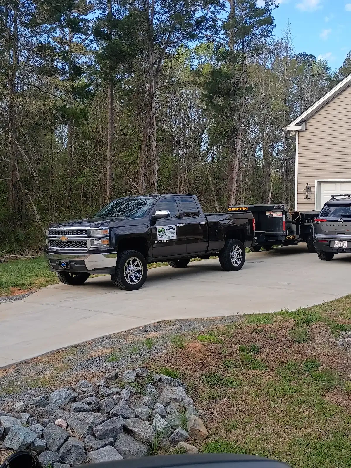 A black Chevrolet Silverado pickup truck parked on a concrete driveway, with a utility trailer attached behind it, next to a beige house with a white garage door, in a residential area with trees in the background.