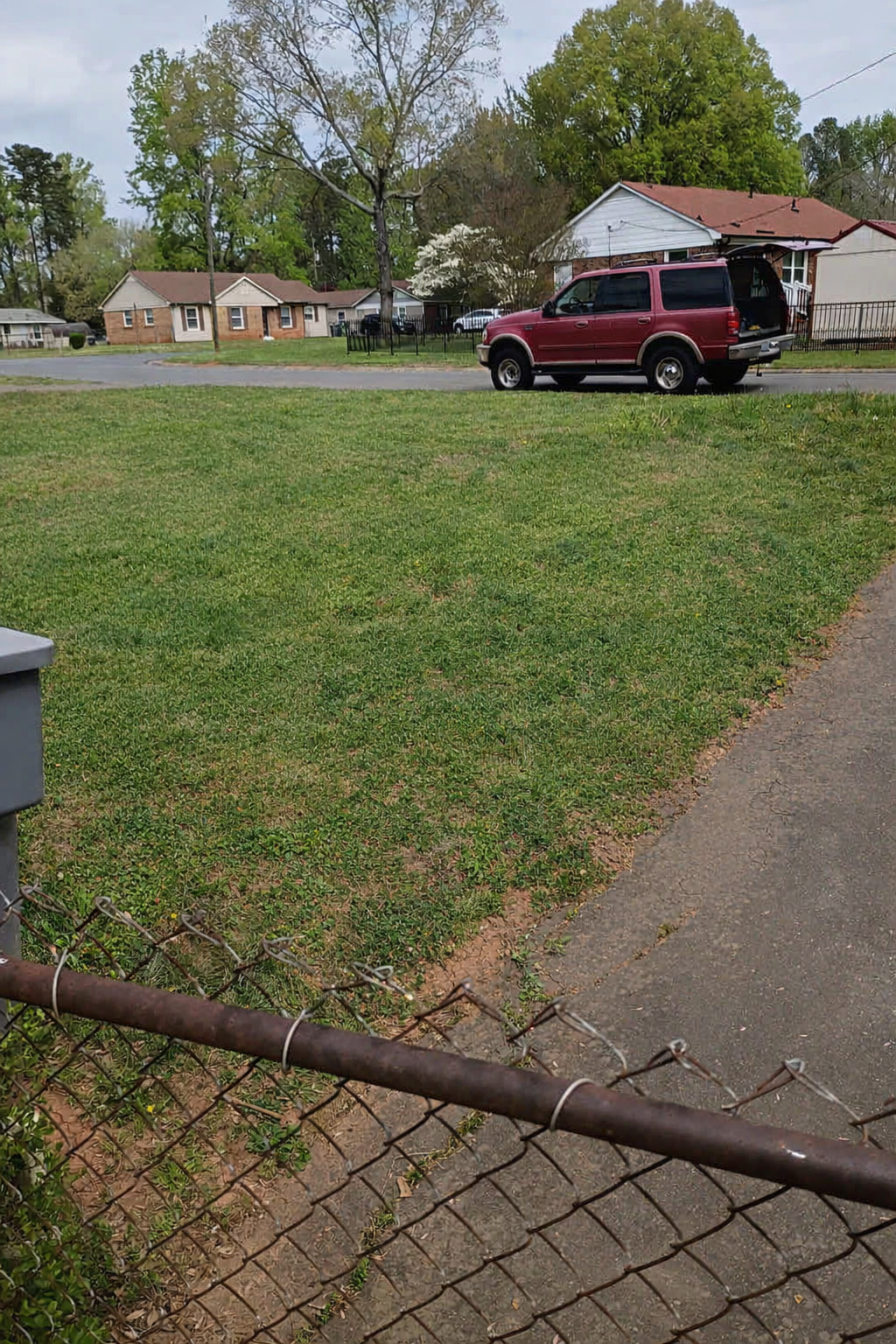 A suburban neighborhood with green lawns, houses, and trees. A red SUV is parked on the street with its rear hatch open. The scene includes a chain-link fence in the foreground and cloudy skies above after Hallelujah Hauling cleaned it.