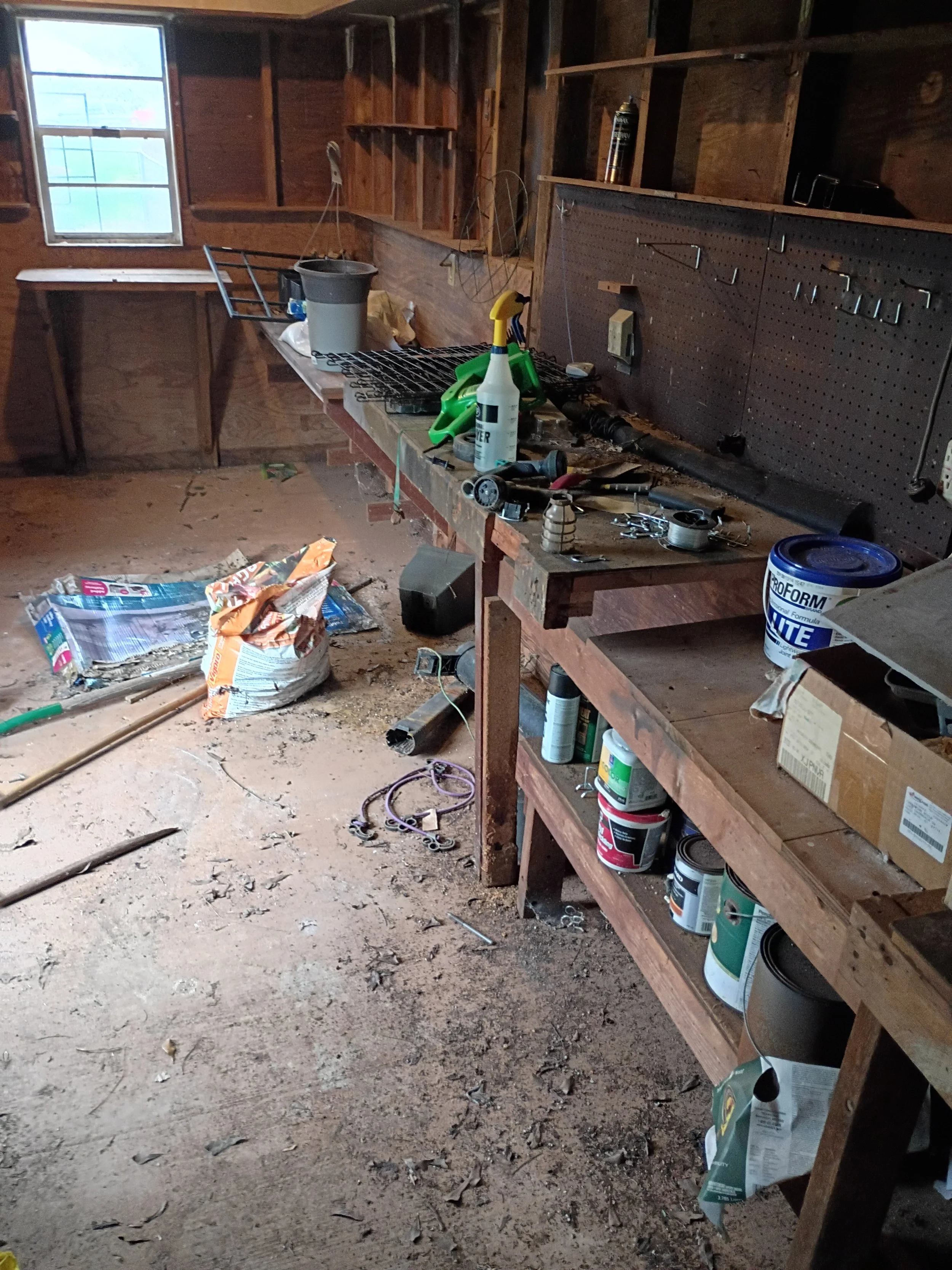 A cluttered garage with a workbench, various tools, paint cans, and supplies. The floor is dusty with scattered debris and the wall has pegs and shelves for storage before Hallelujah Hauling cleaned it.