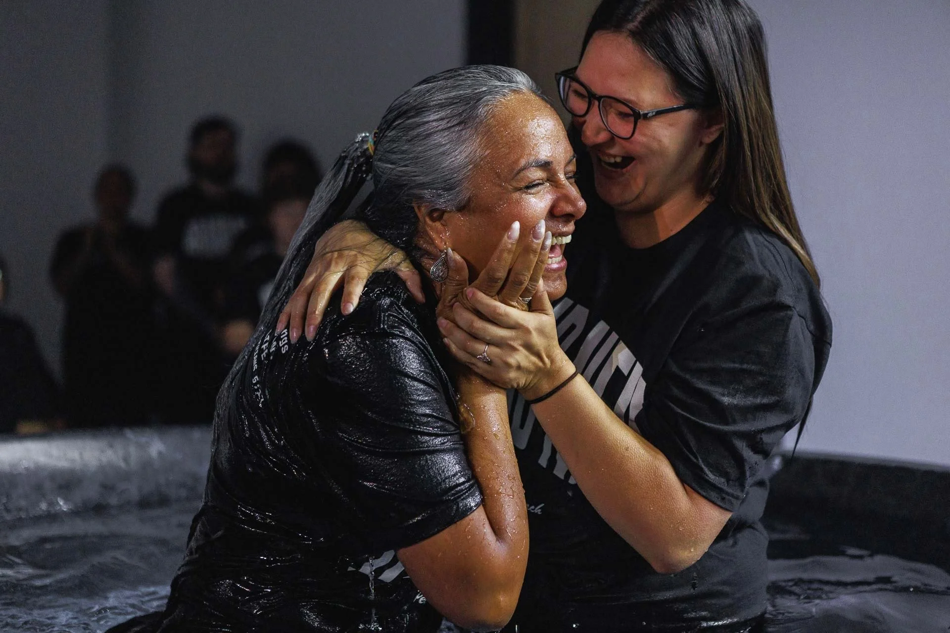 Two women sharing an emotional moment in water; one woman with grey hair and dark wet clothing, the other woman with glasses and brown hair, embracing and smiling.