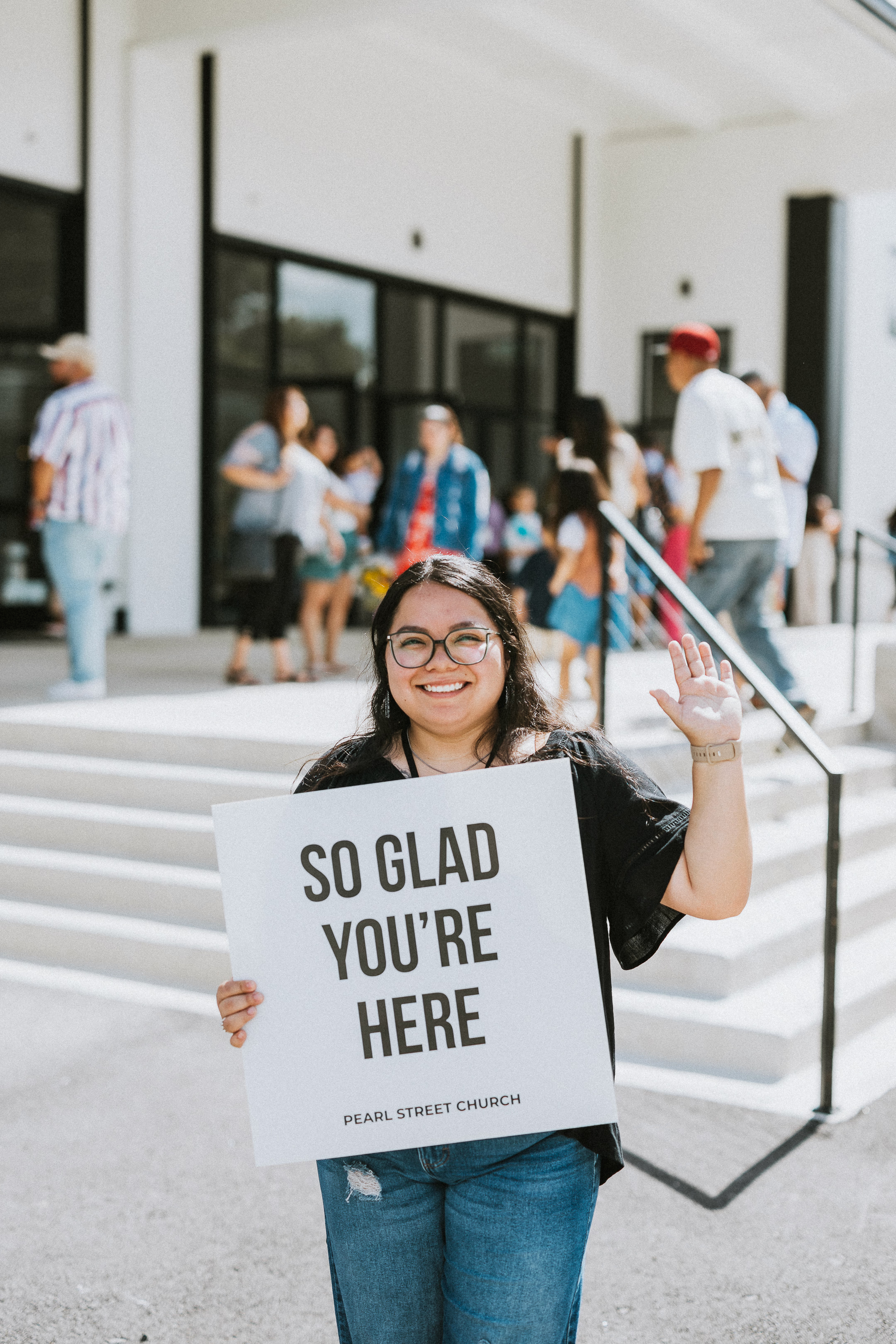 Welcome Team host holding a "So Glad You're Here" sign outside Pearl Street Church Downtown