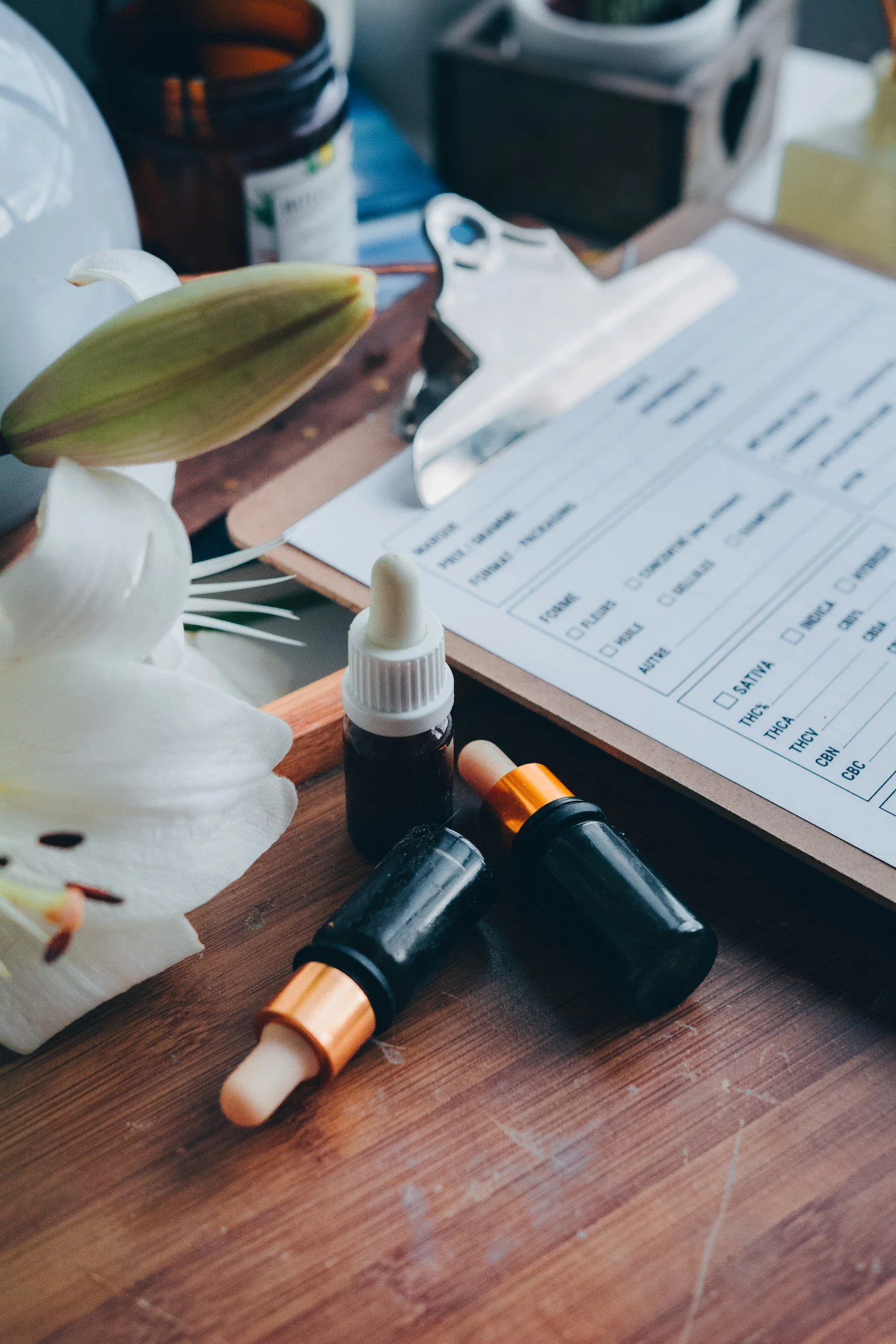 A wooden table with cannabis oil bottles, a syringe, a clipboard with a medical form, and a white orchid flower.