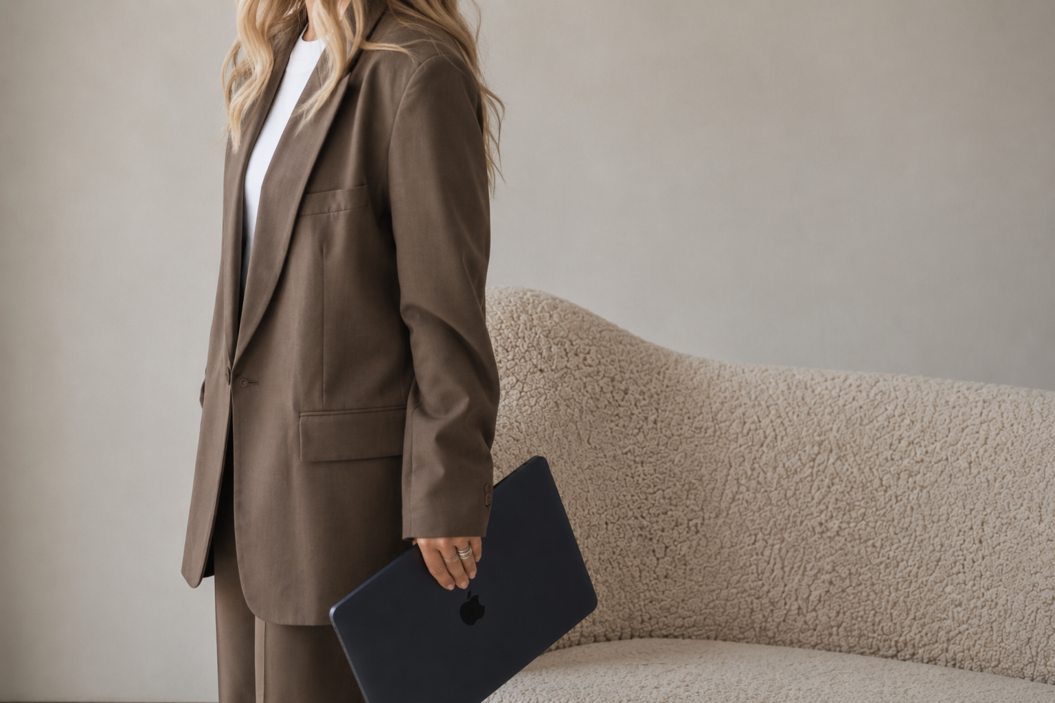 A woman in a brown suit holding a closed dark gray Apple MacBook, standing next to a beige textured sofa.
