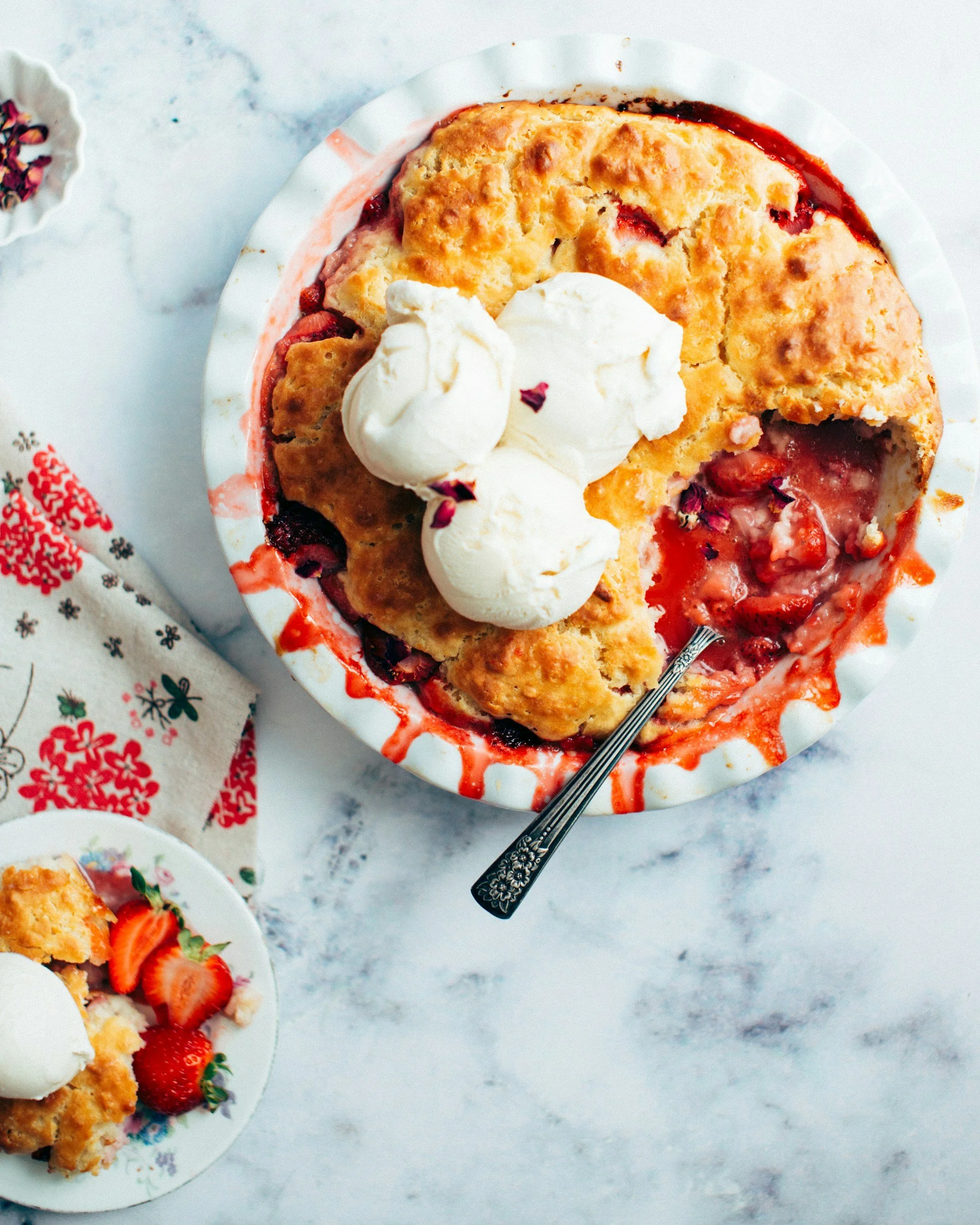 A slice of strawberry rhubarb pie topped with whipped cream served in a paper dish on a white marble surface, with a decorative fork.
